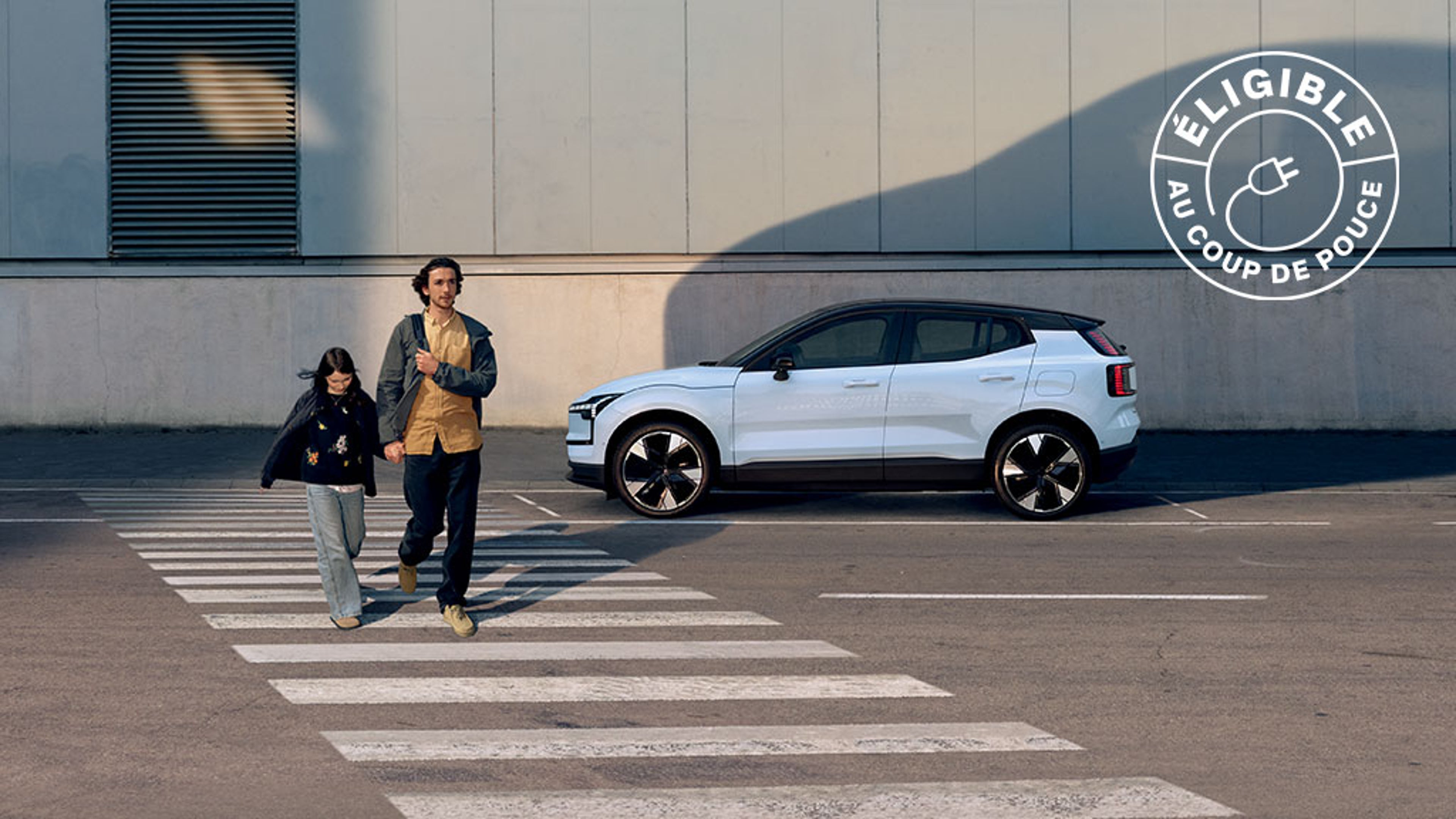 Deux personnes traversent un passage piéton, Volvo EX30 blanche en arrière-plan projetant une ombre sur le mur