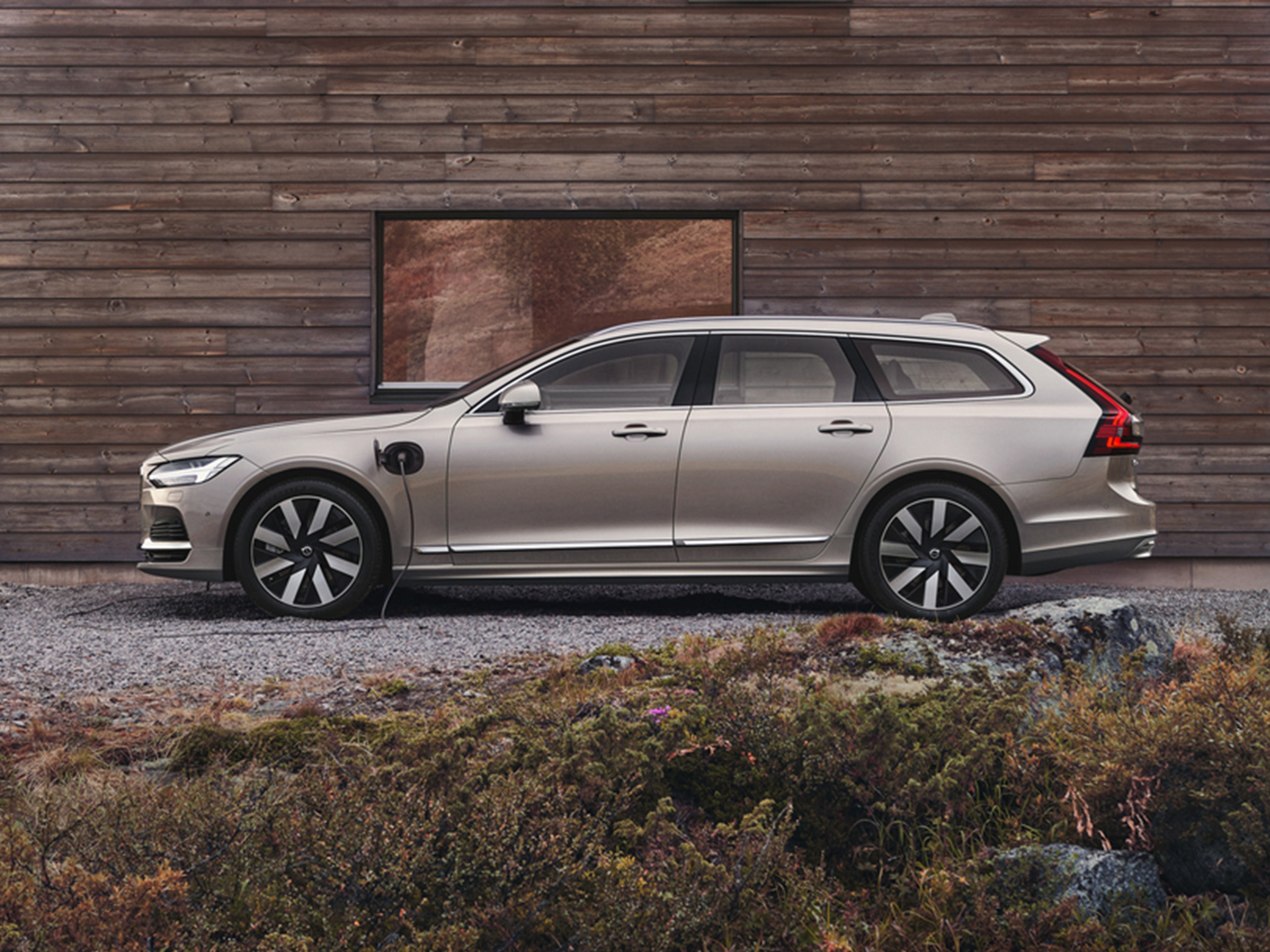 Bright Dush -värinen Volvo V90 lataushybridifarmari auto pysäköitynä pihalle puisen talon seinän eteen.