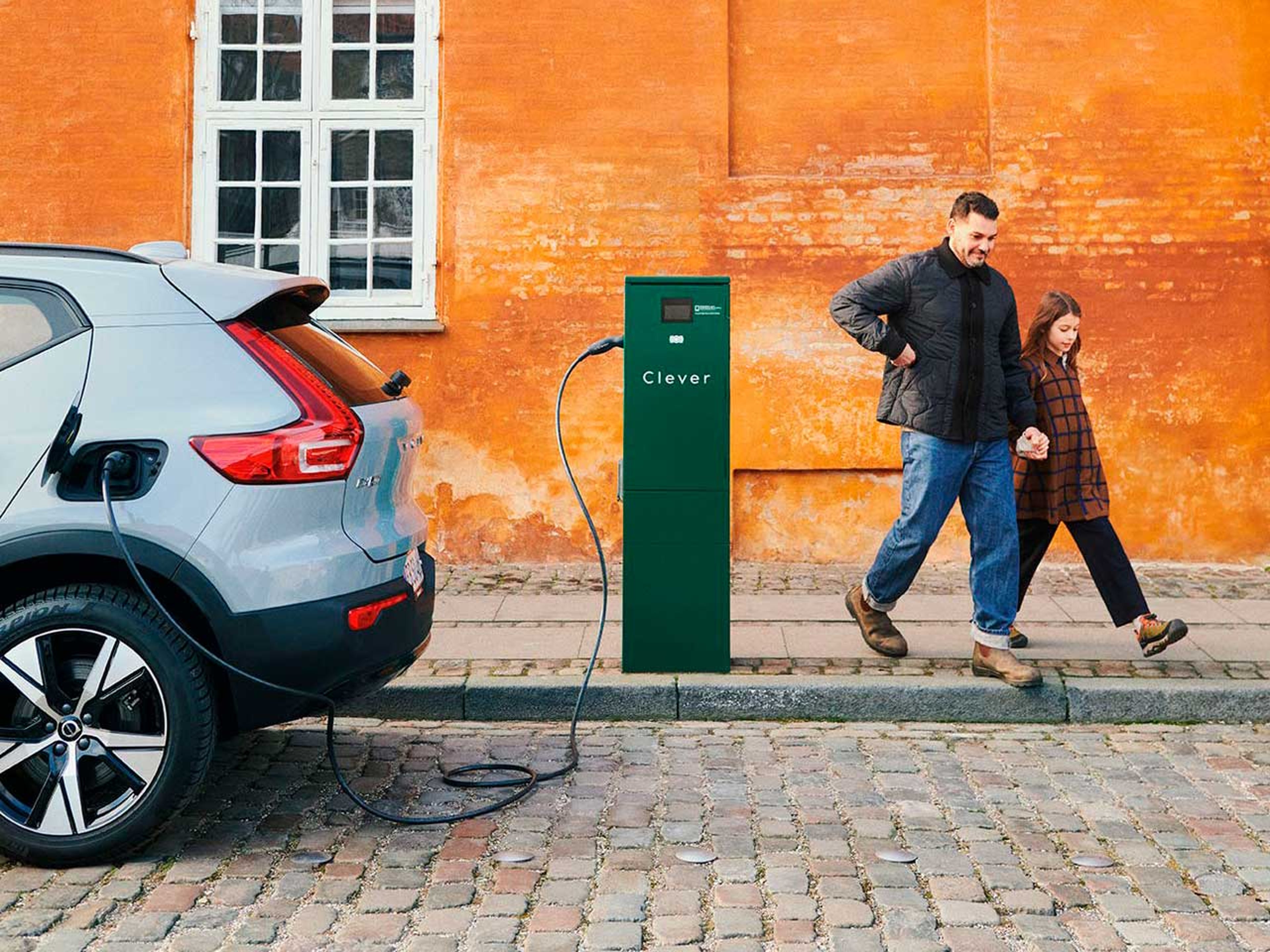 En mand og en pige går hånd i hånd ved siden af en Volvo elbil, der lader op ved en Clever-ladestation. Baggrunden viser en orange væg og et vindue.