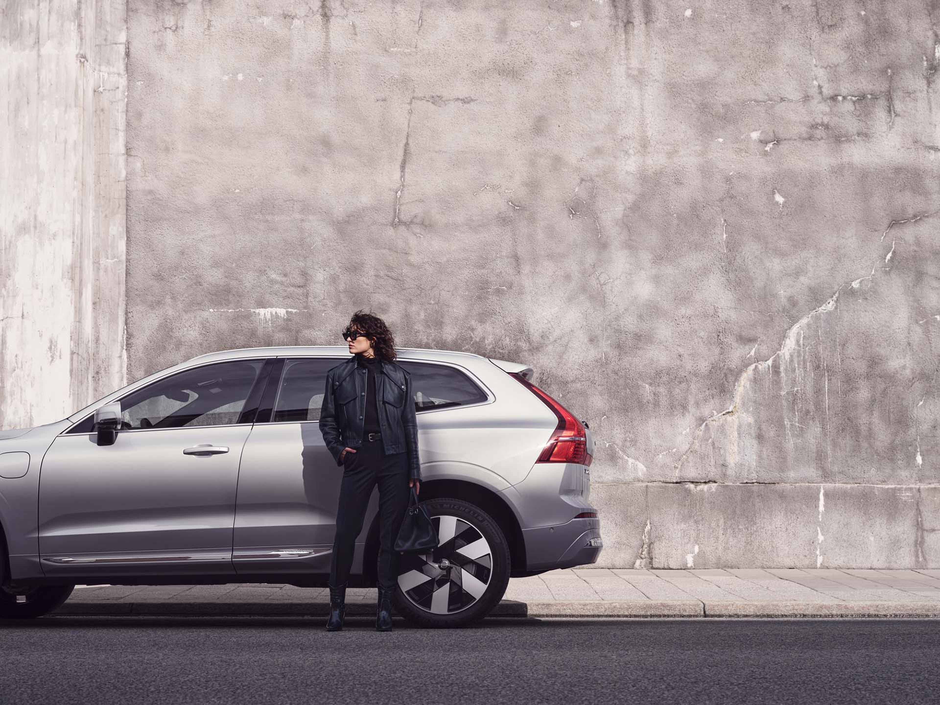 Volvo XC60 de perfil, junto a uma parede cinzenta de betão, junto a um passeio, com uma mulher vestida de preto encostada à porta traseira a olhar para o lado esquerdo