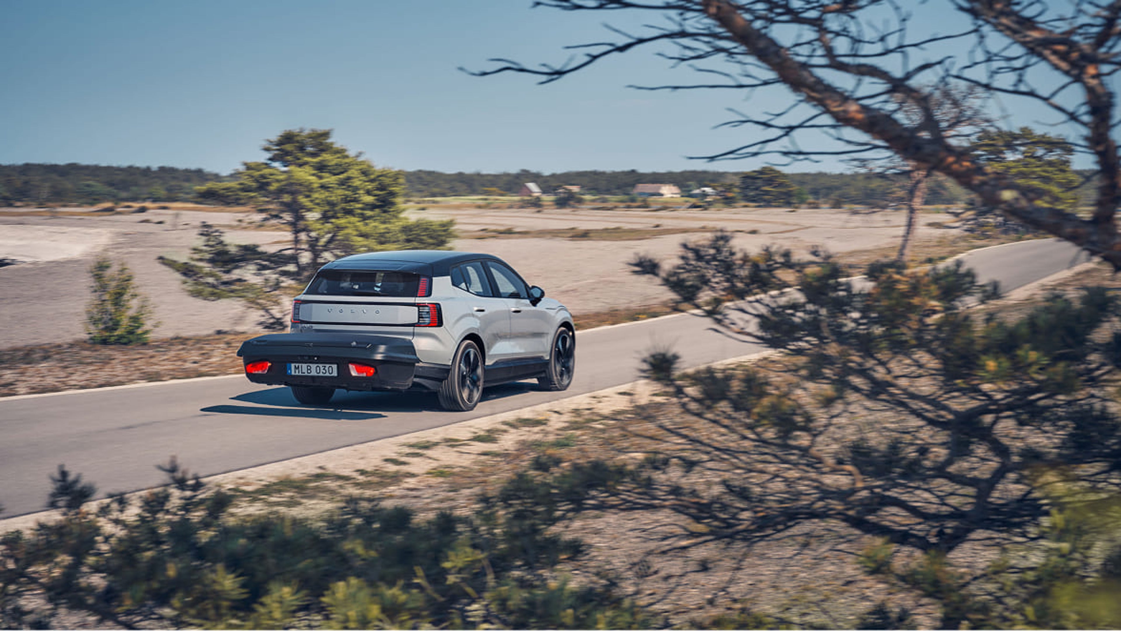 Volvo EX30 visto por trás com uma caixa de carga montada na barra de reboque, a conduzir numa estrada rural