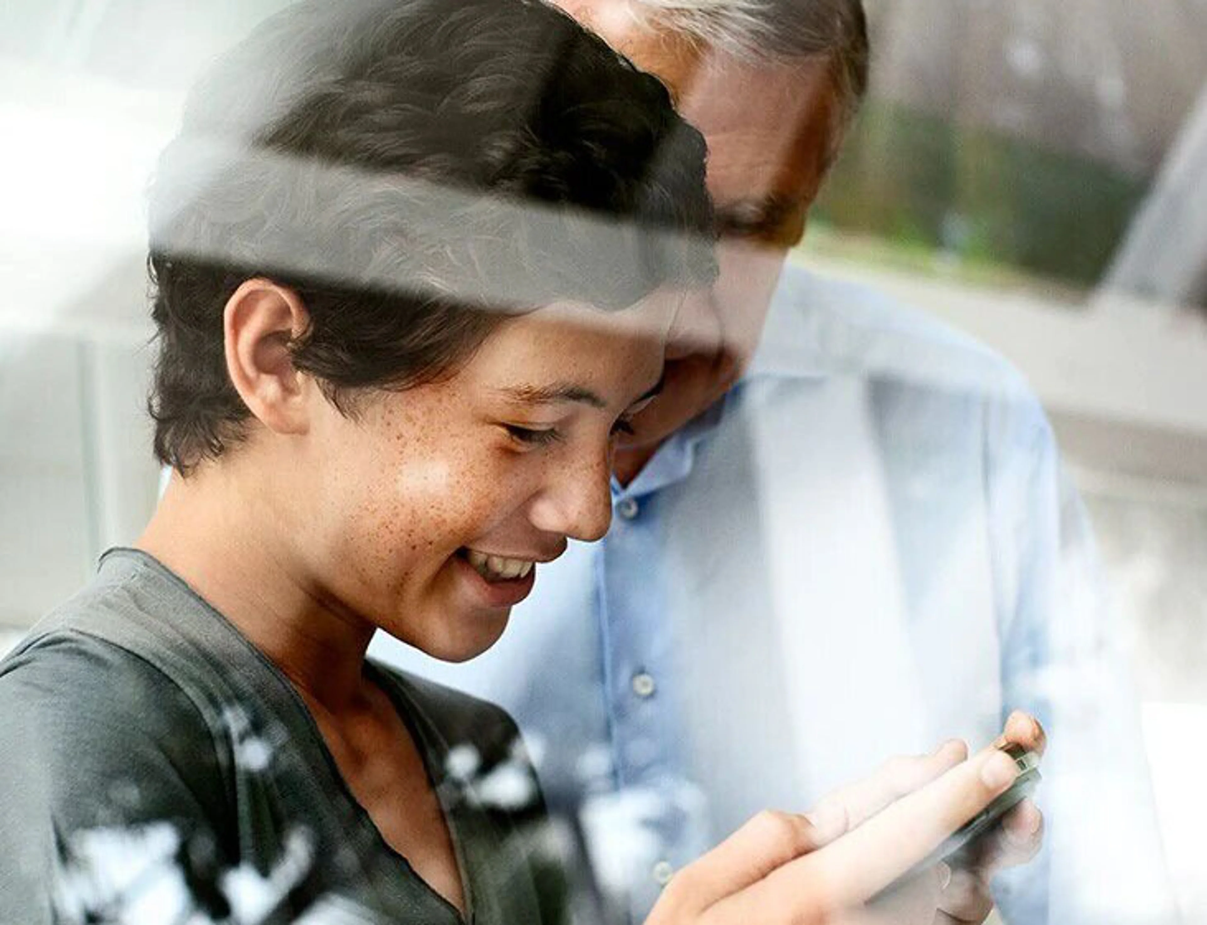 Une jeune personne regarde un smartphone en souriant, tandis qu'une personne plus âgée regarde par-dessus son épaule à l'arrière-plan. L'image présente des reflets, comme si elle était prise à travers une vitre.