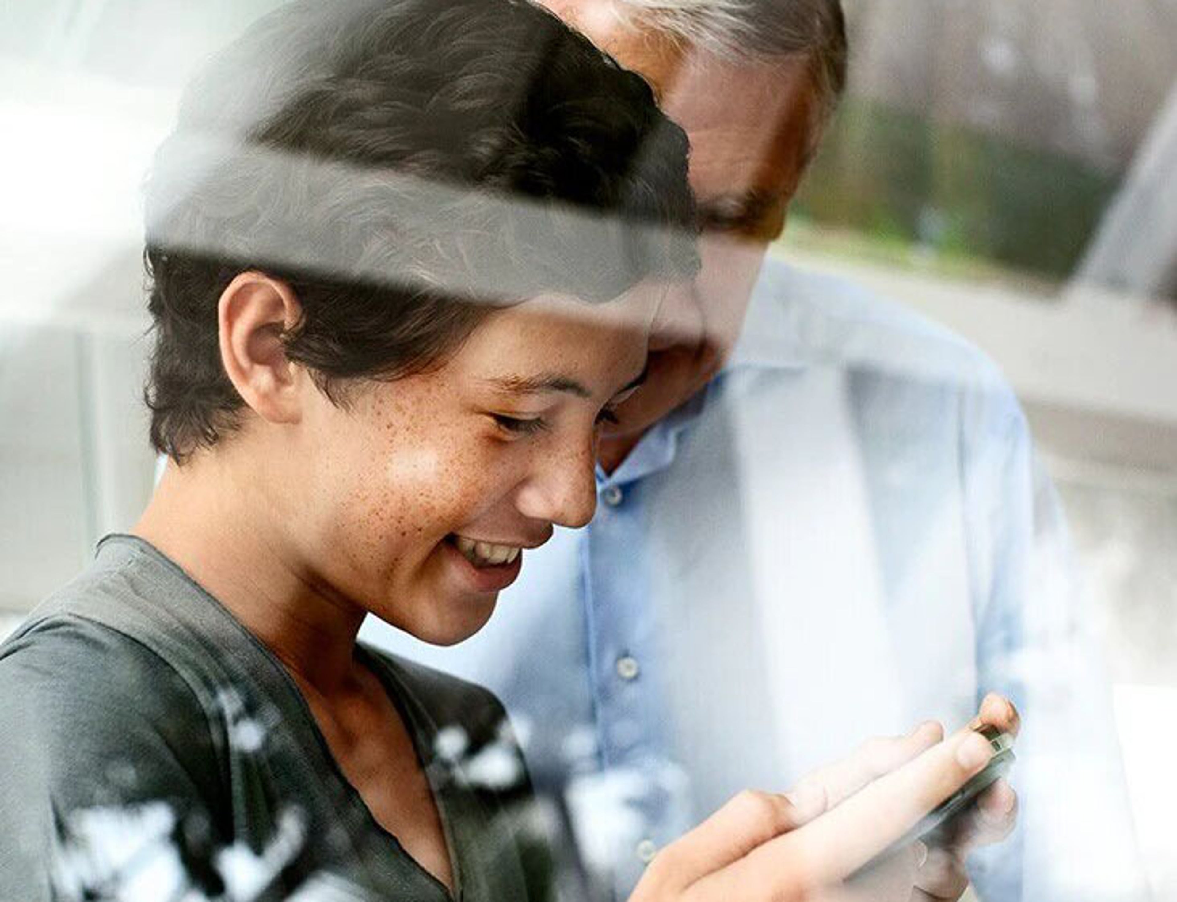 Une jeune personne regarde un smartphone en souriant, tandis qu'une personne plus âgée regarde par-dessus son épaule à l'arrière-plan. L'image présente des reflets, comme si elle était prise à travers une vitre.