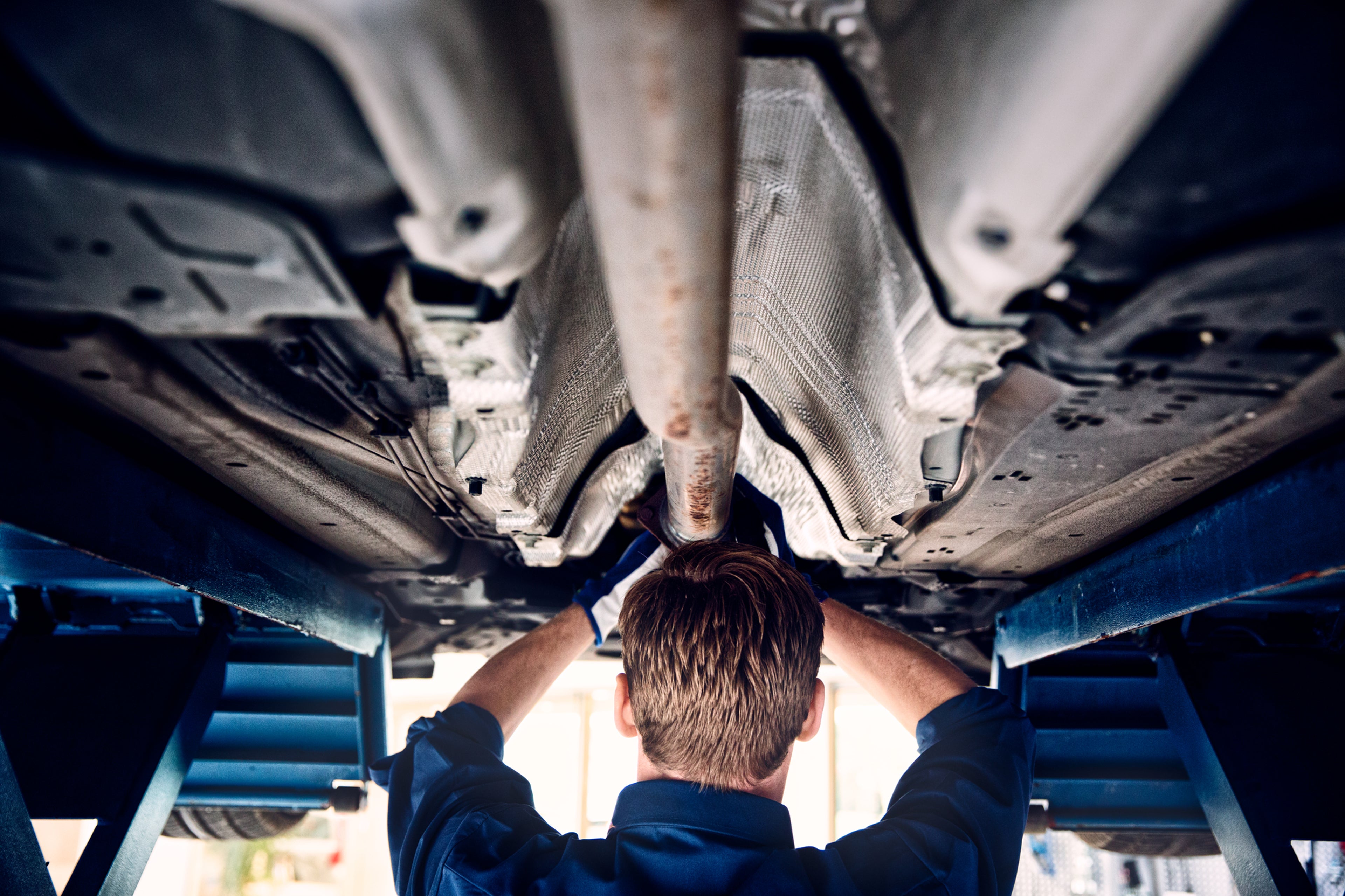 Снимка на мъж в гръб, облечен в синя риза, който извършва ремонт под автомобил, вдигнат над него, докато се намира в сервизен тунел.