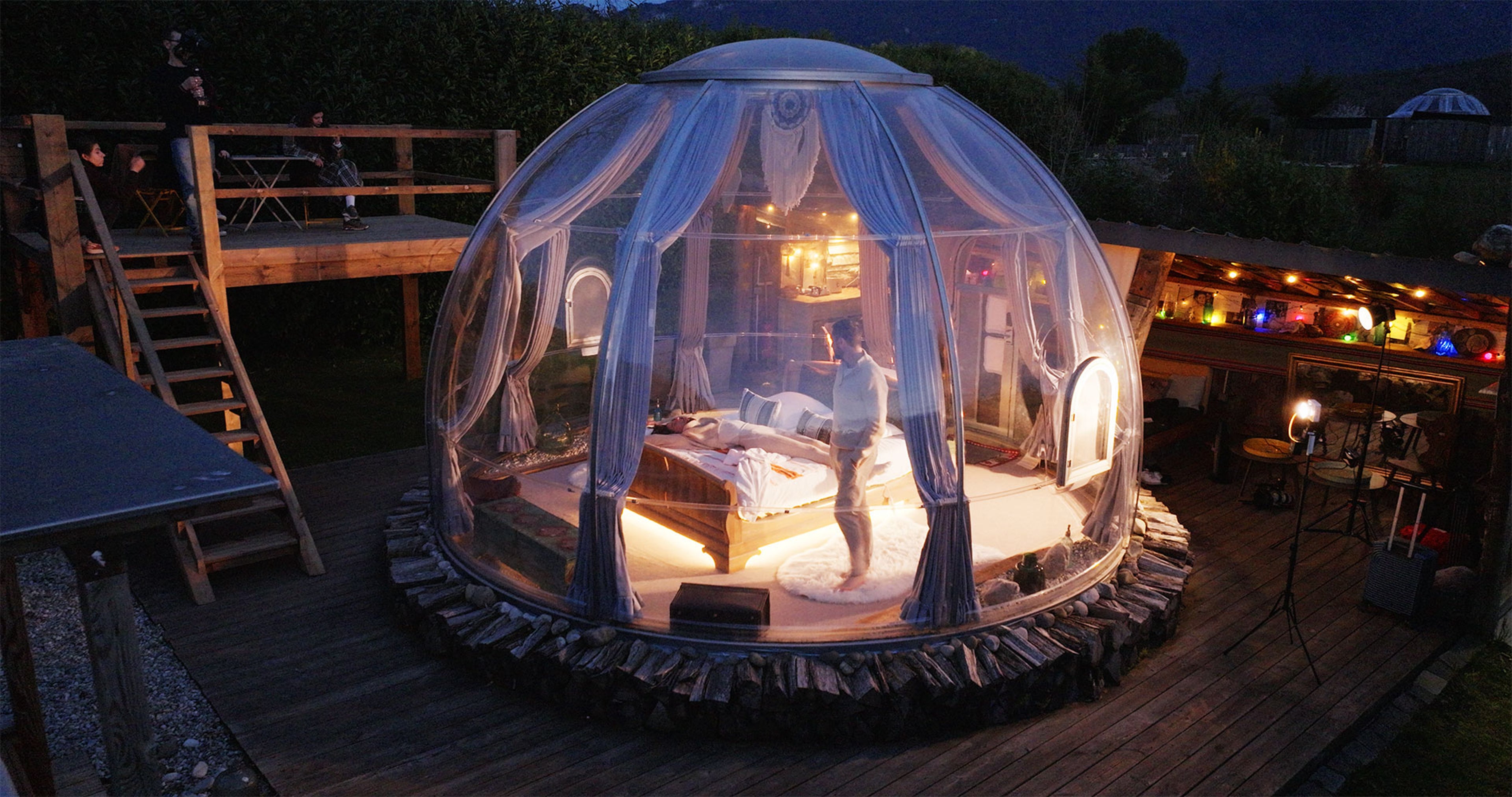 Dôme transparent éclairé avec lit et rideaux sur terrasse en bois, personnes visibles à l’extérieur.