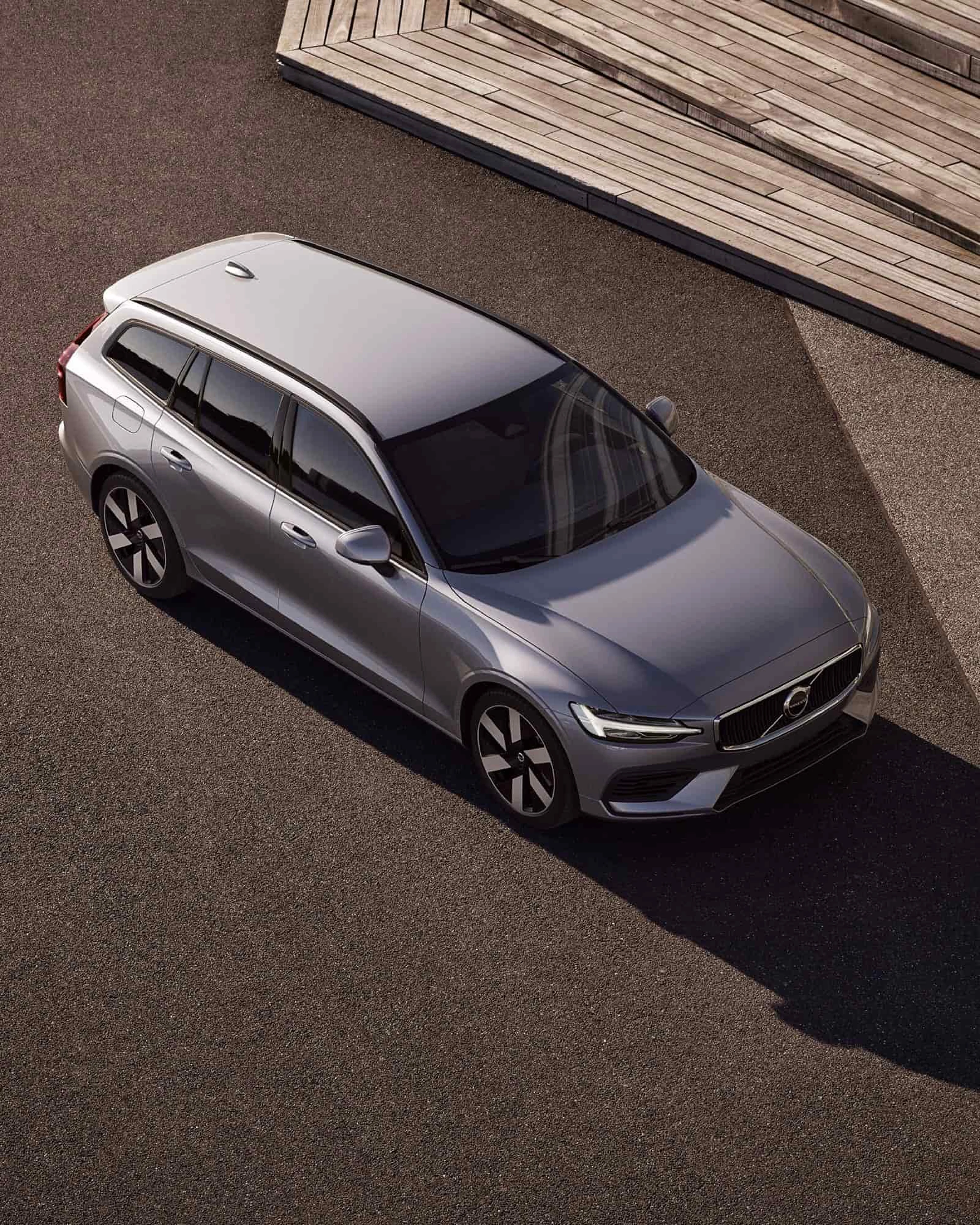 Vue aérienne d'une Volvo V60 hybride rechargeable grise roulant sur un chemin de terre avec un escalier en bois sur le côté.