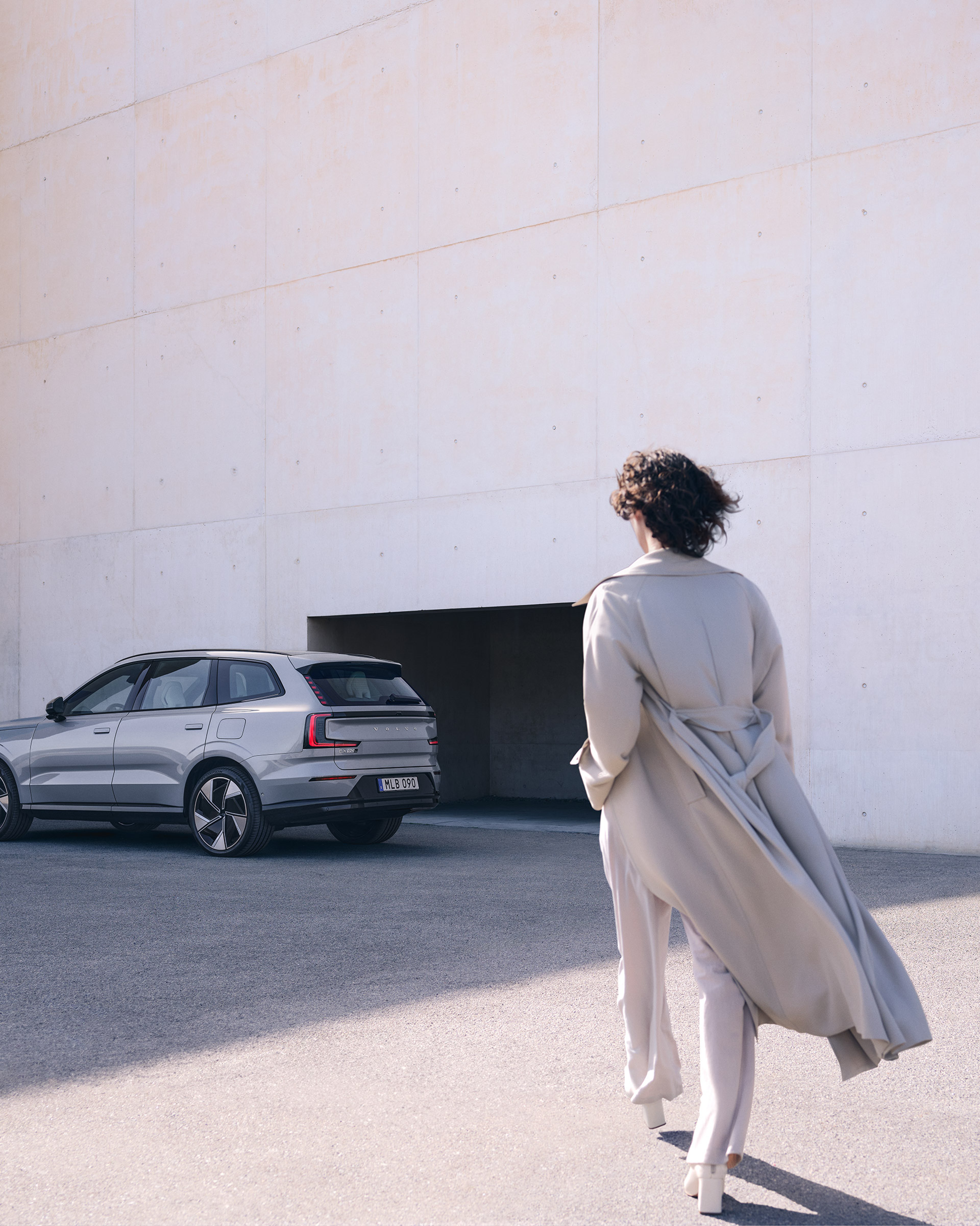 Vaaleaan trenssiin pukeutunut nainen kävelee kohti Vapour Grey -väristä Volvo EX90 täyssähköautoa pysäköintialueella.
