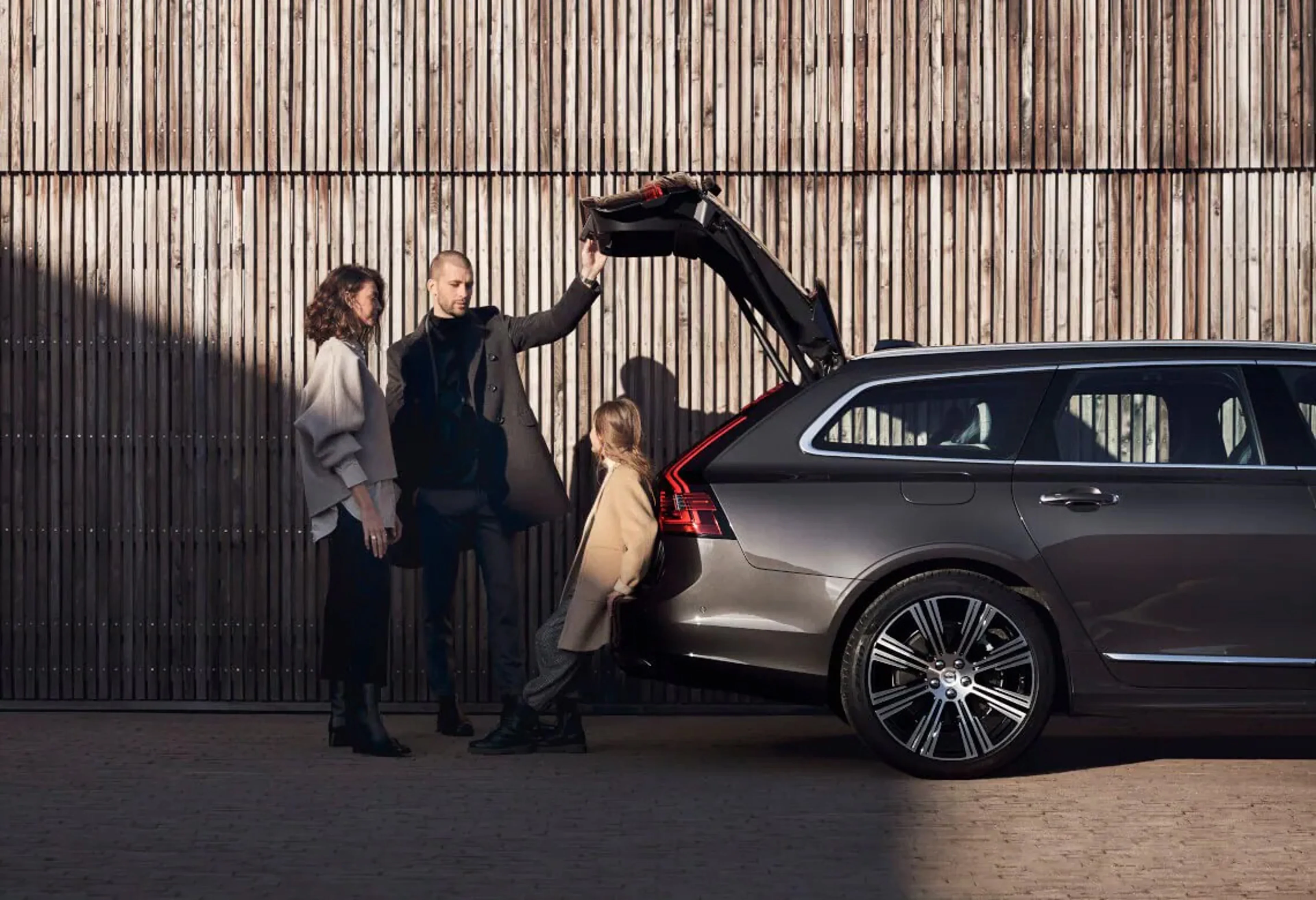 Eine dreiköpfige Familie steht an der geöffneten Heckklappe eines anthrazitfarbenen Volvo V90. Der Mann (flankiert von einer Frau) hält die Klappe geöffnet, während die Tochter sich rückwärtig gegen das Fahrzeug lehnt.
