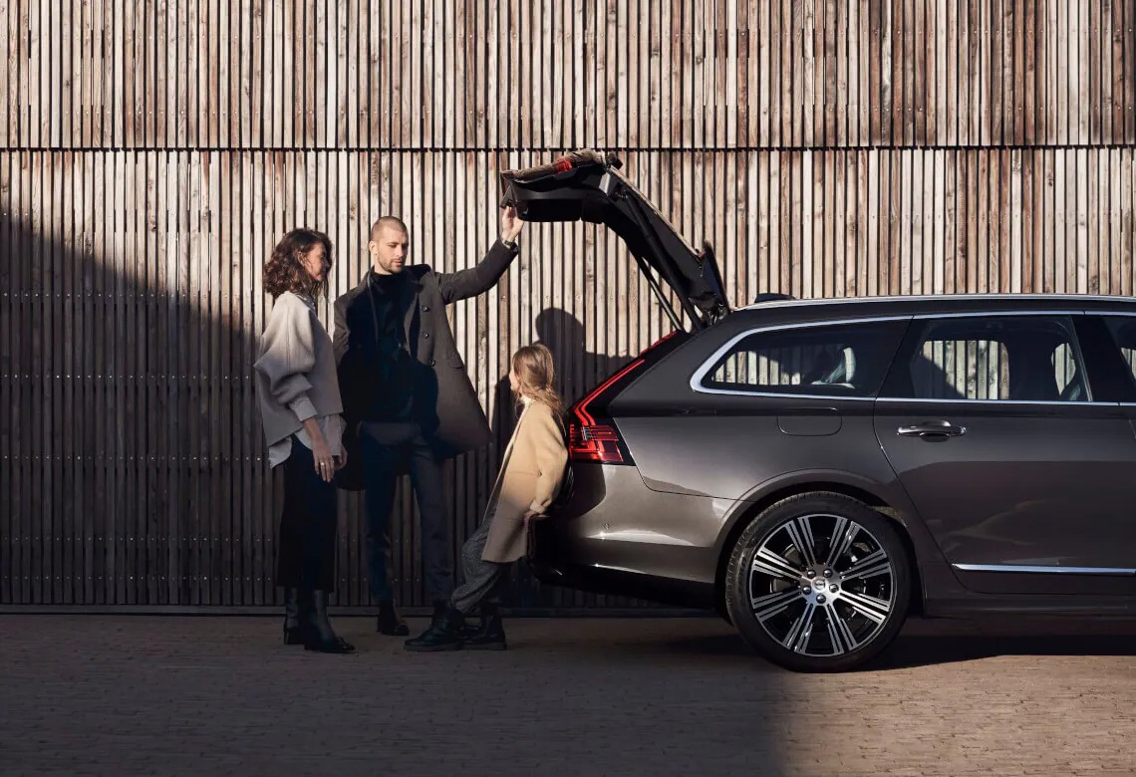 Eine dreiköpfige Familie steht an der geöffneten Heckklappe eines anthrazitfarbenen Volvo V90. Der Mann (flankiert von einer Frau) hält die Klappe geöffnet, während die Tochter sich rückwärtig gegen das Fahrzeug lehnt.