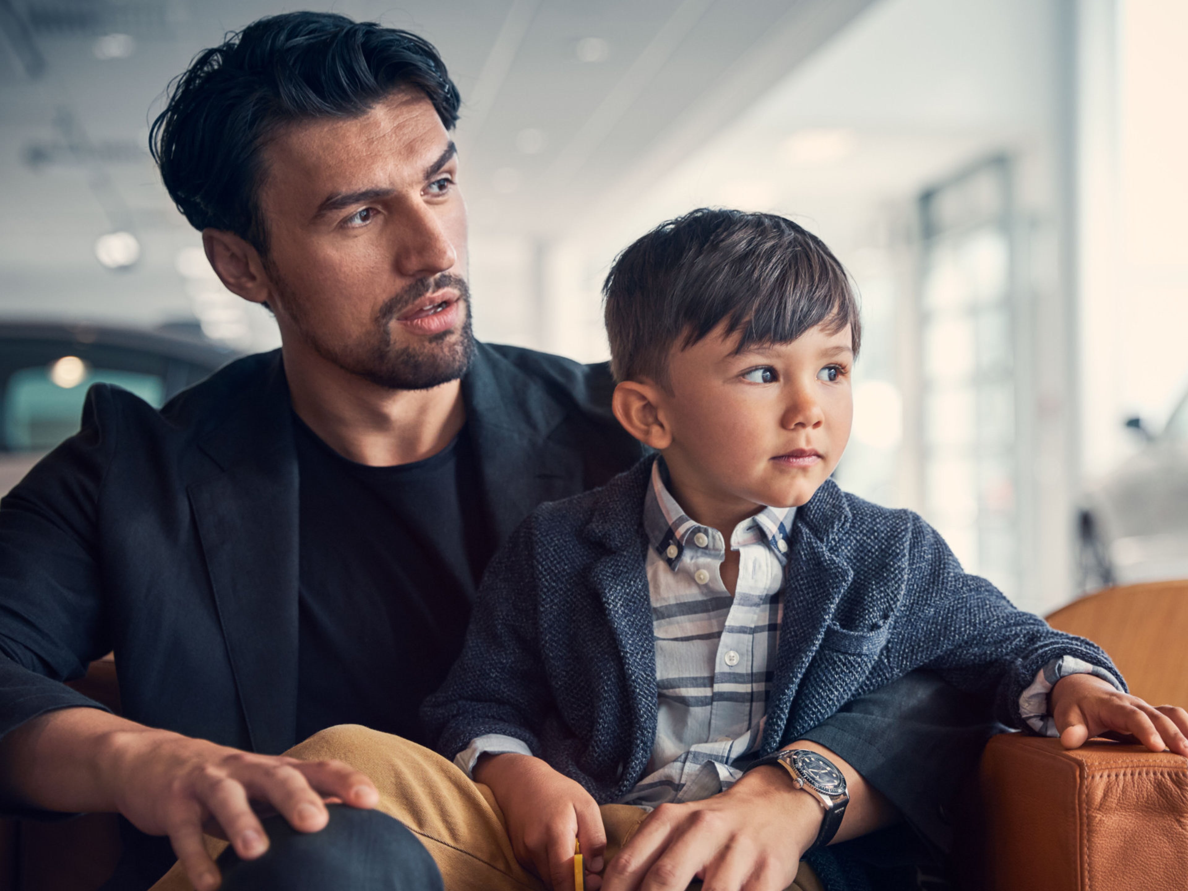 Ein dunkelhaariger Mann hat seinen Arm um seinen Sohn gelegt. Beide tragen einen Anzug und schauen in Richtung Volvo Werkstatt.