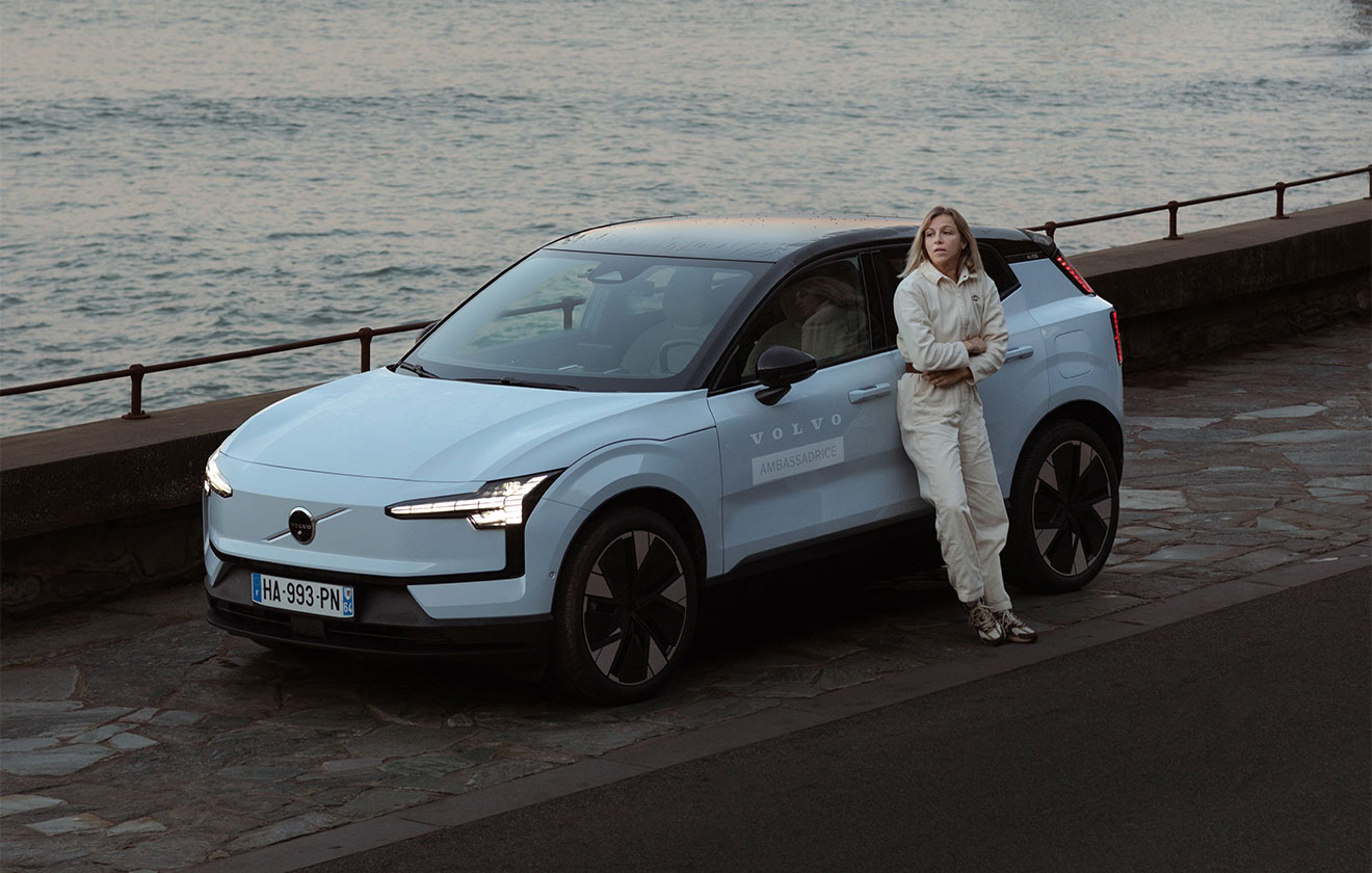 Volvo EX30 bleu nuage stationnée en bord de mer avec Julie Bourges en appuyée contre la voiture