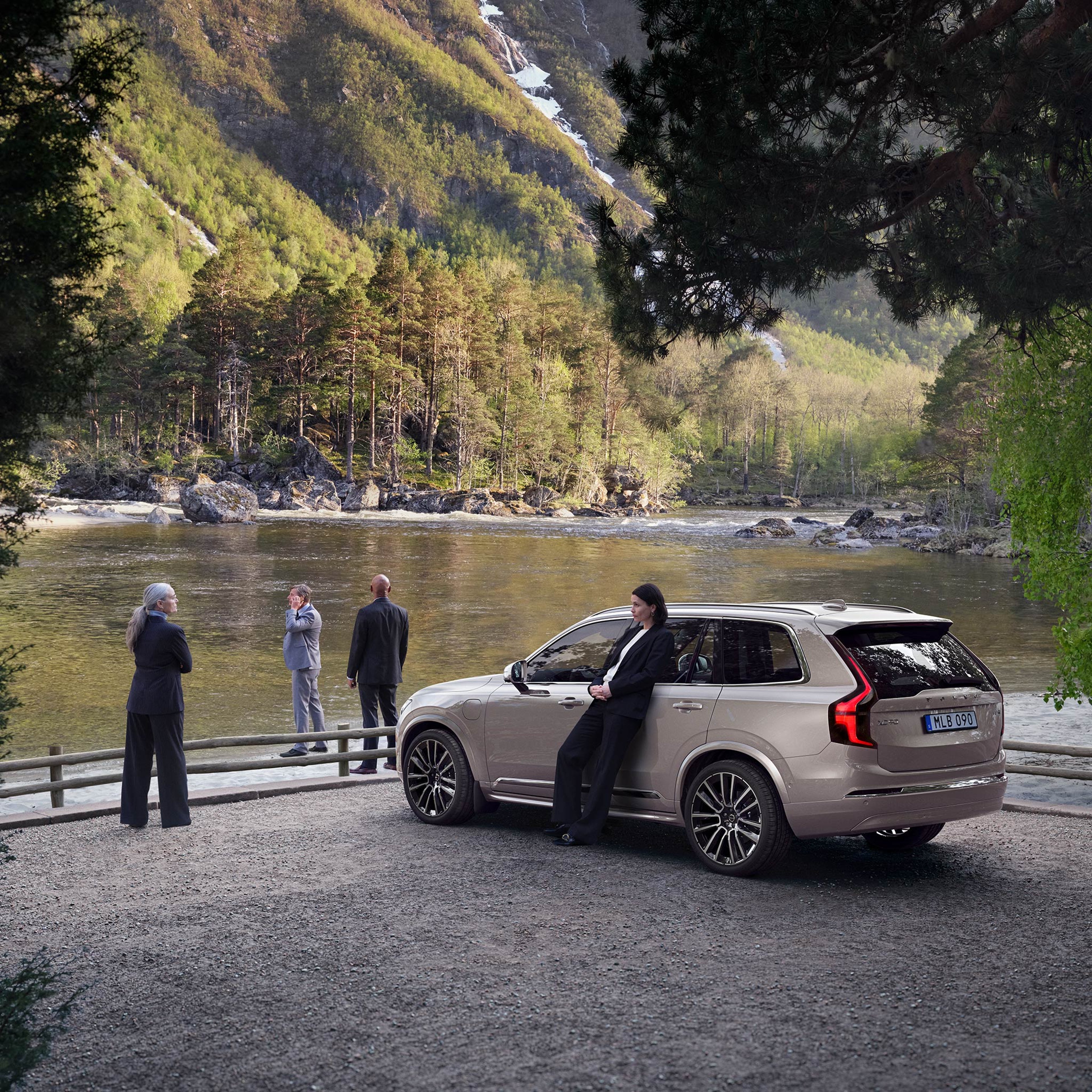 Mustaan housupukuun pukeutunut nainen nojaa joen rantaan parkkeerattuun Bright Dusk -väriseen Volvo XC90 lataushybridikatumaasturin. Auton lähettyvillä, lähempänä rantaa, seisoo mustaan housupukuun pukeutunut nainen, tummaan pukuun pukeutunut mies ja harmaaseen pukuun pukeutunut mies, joka puhuu puhelimessa.