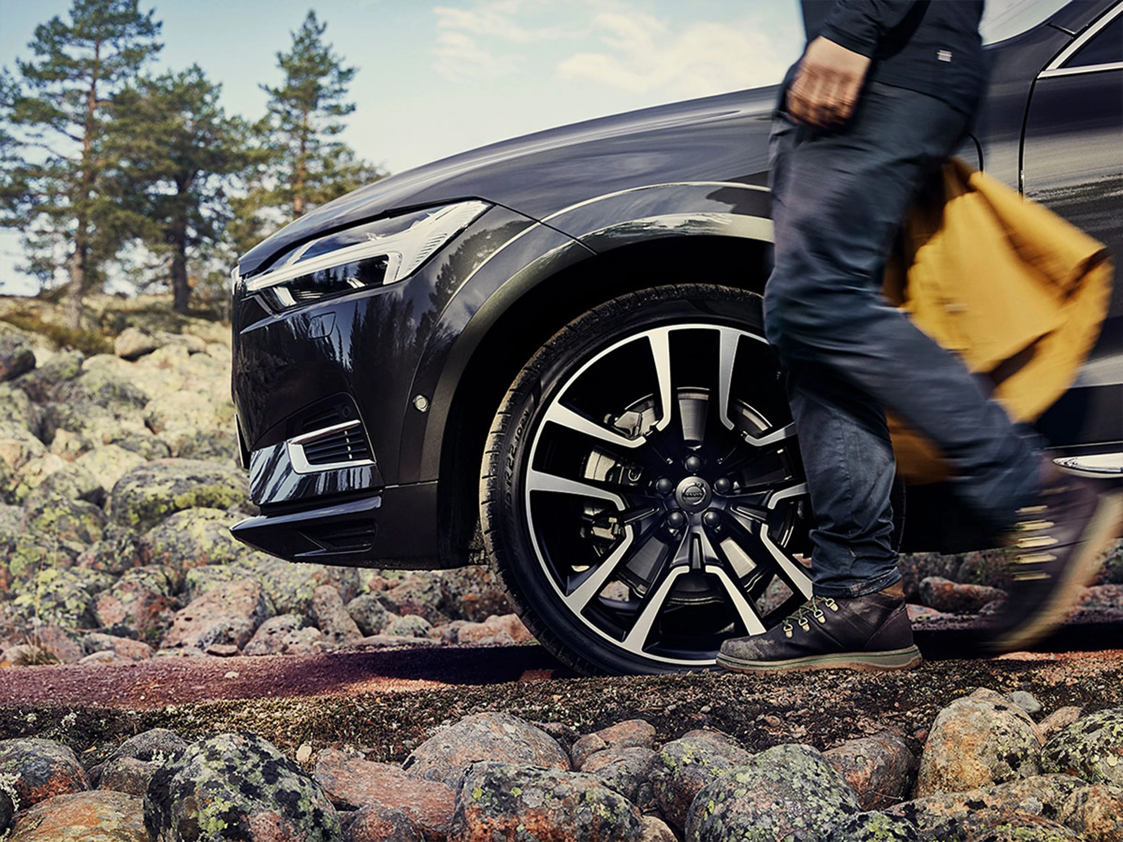 Une Volvo XC60 noire est garée sur un terrain rocheux, tandis qu'une personne, une veste jaune à la main, passe à côté.