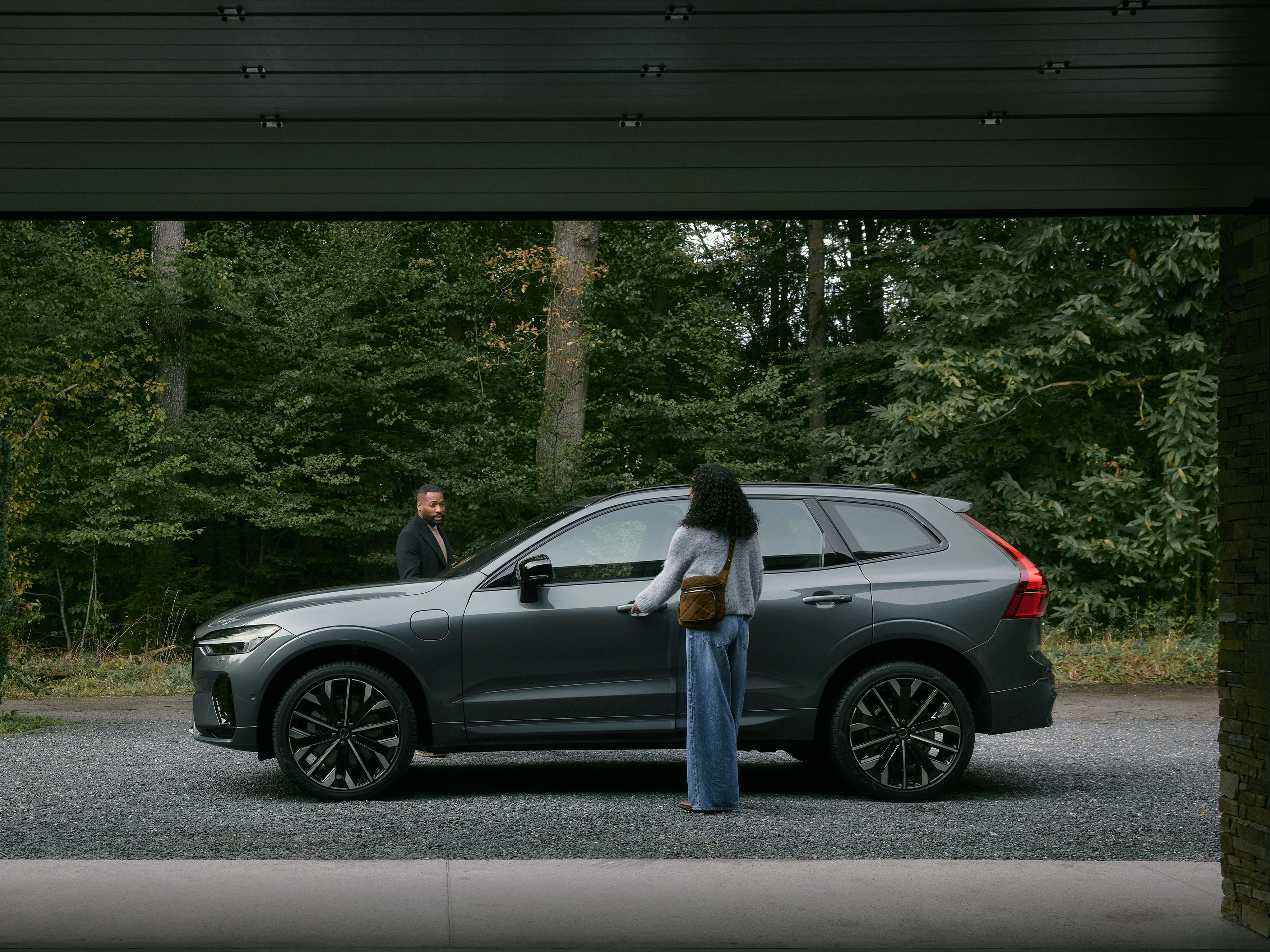Zwei Personen sind dabei in einen dunkelgrauen Volvo XC60 SUV einzusteigen, der vor einem Wald steht.