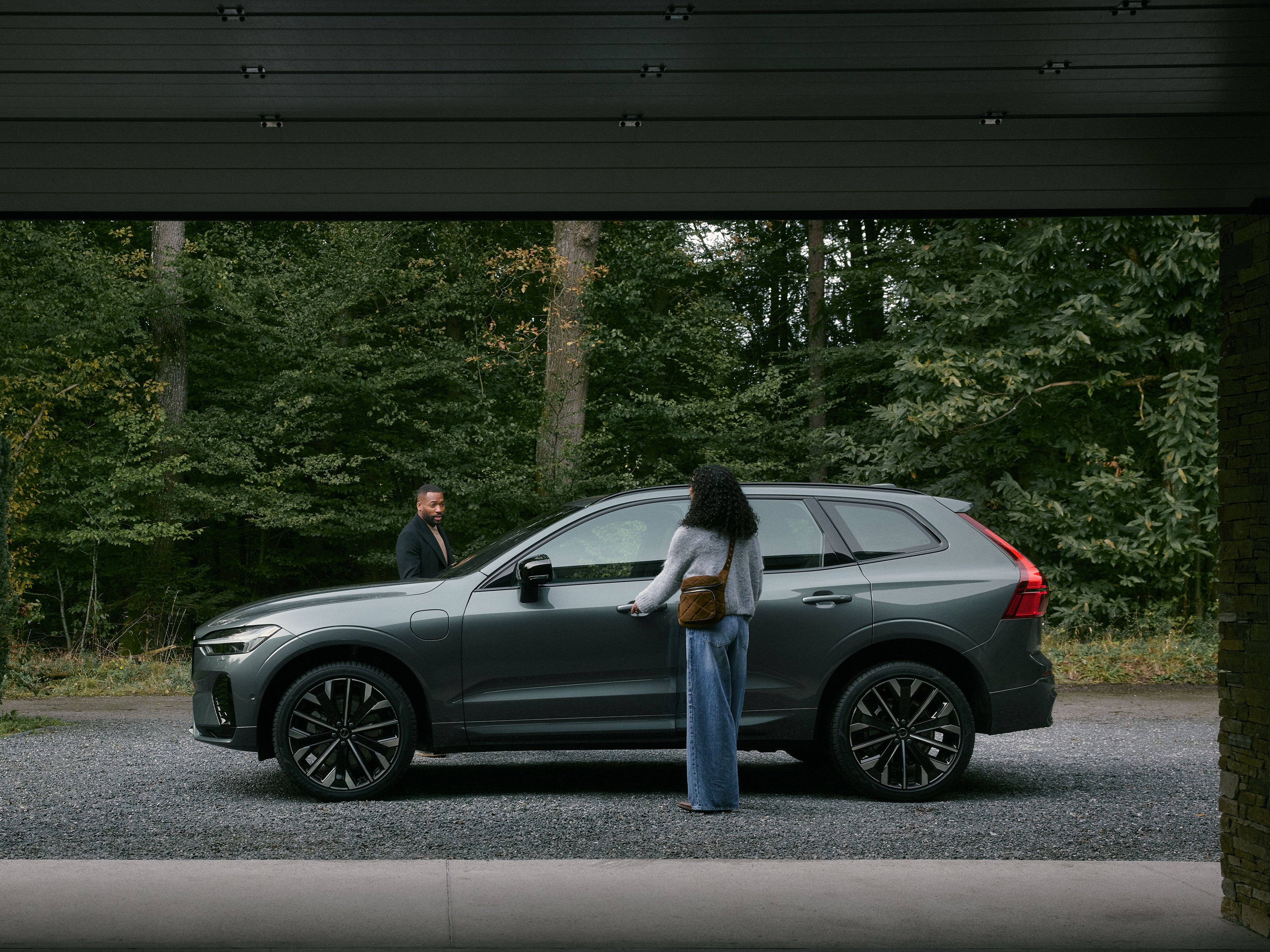 Zwei Personen sind dabei in einen dunkelgrauen Volvo XC60 SUV einzusteigen, der vor einem Wald steht.