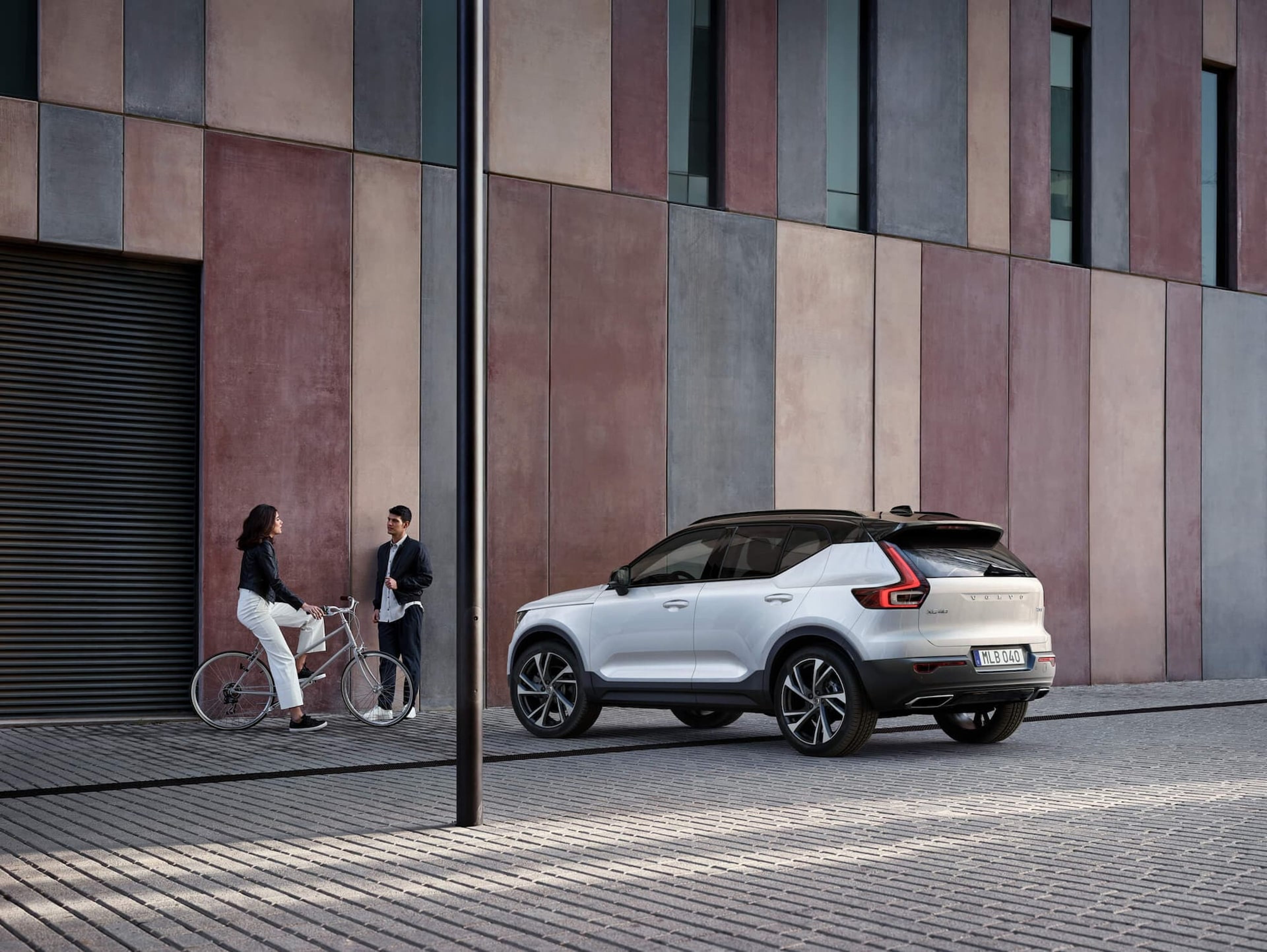 Ein Mädchen sitzt auf einem Fahrrad und lehnt an einer Wand, während sie sich mit einem Jungen unterhält. Beide stehen vor einem weißen Volvo XC40 SUV mit schwarzem Dach.
