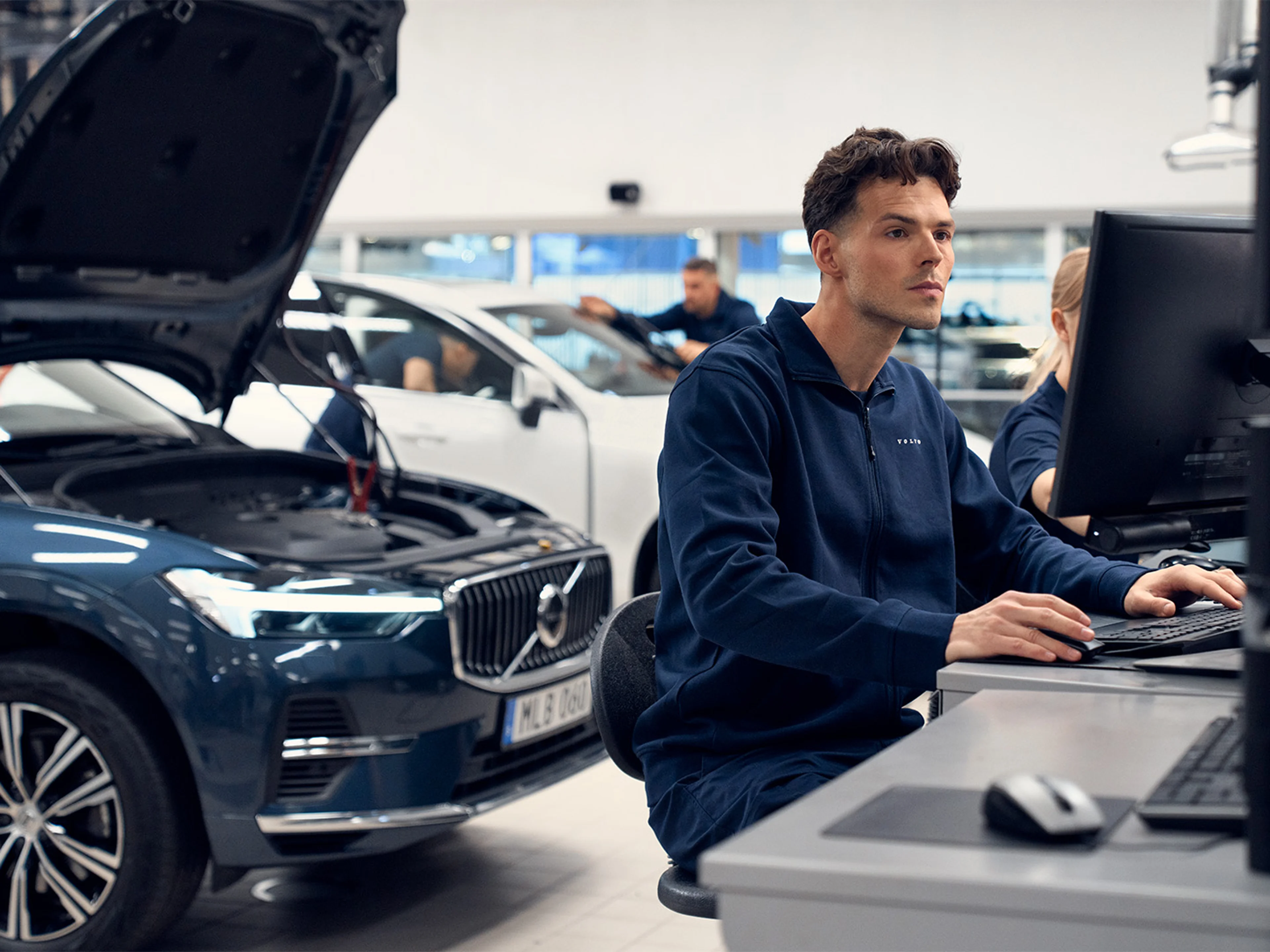 A technician works on a computer in a modern garage, focused on diagnostics, with a Volvo car's engine visible nearby.