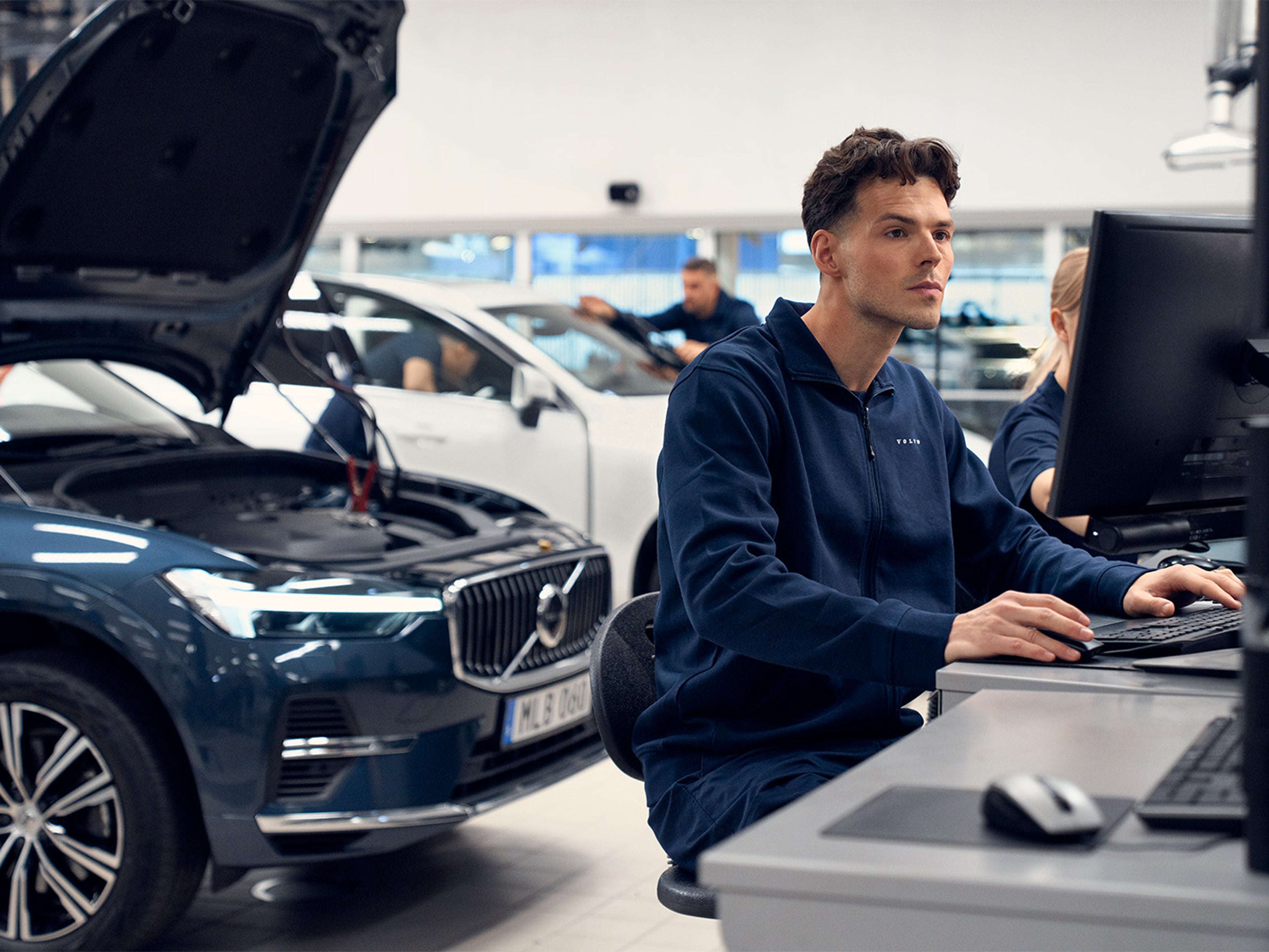 A technician works on a computer in a modern garage, focused on diagnostics, with a Volvo car's engine visible nearby.