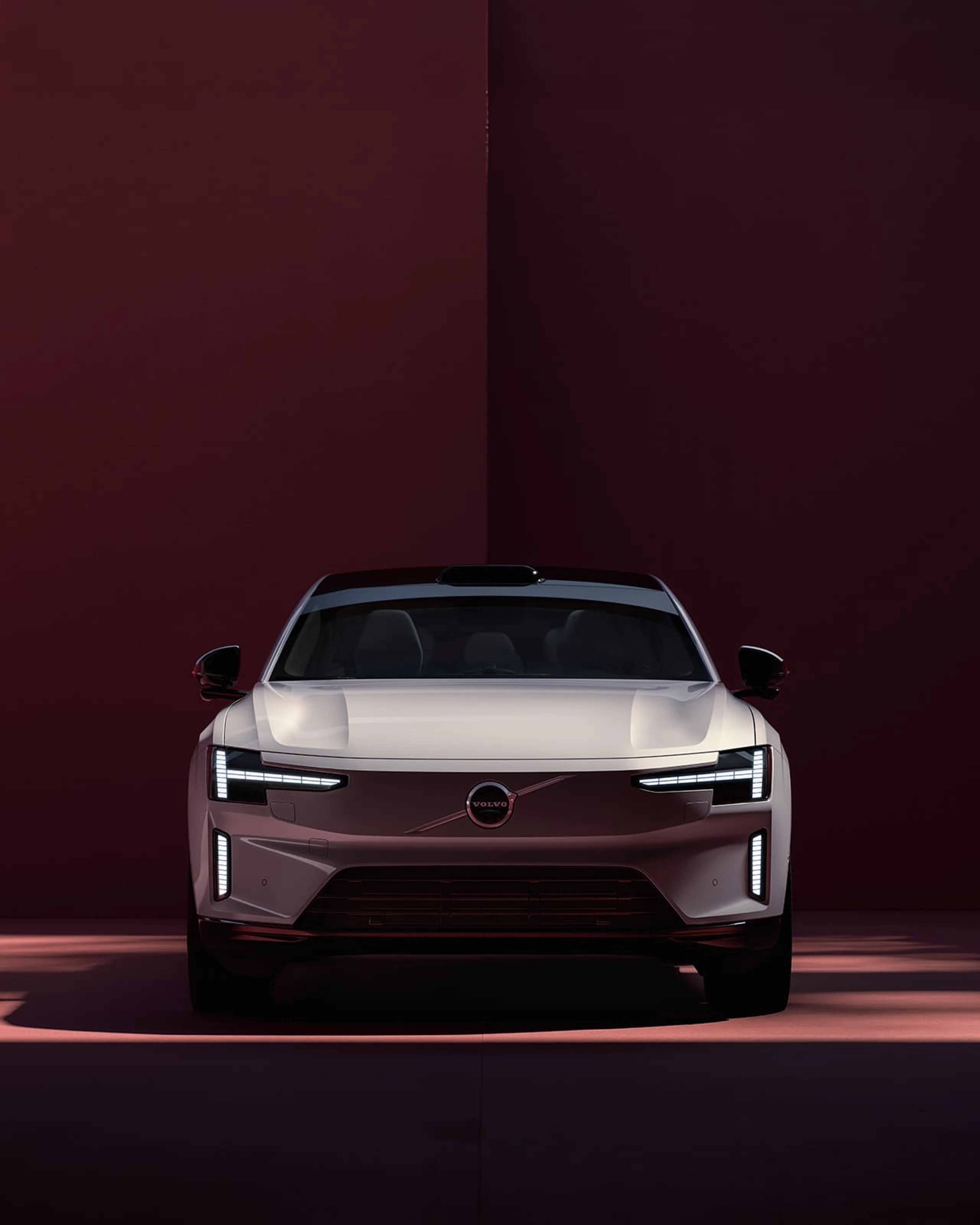 Vooraanzicht van een grijze Volvo ES90 op een bordeauxrode studioachtergrond.