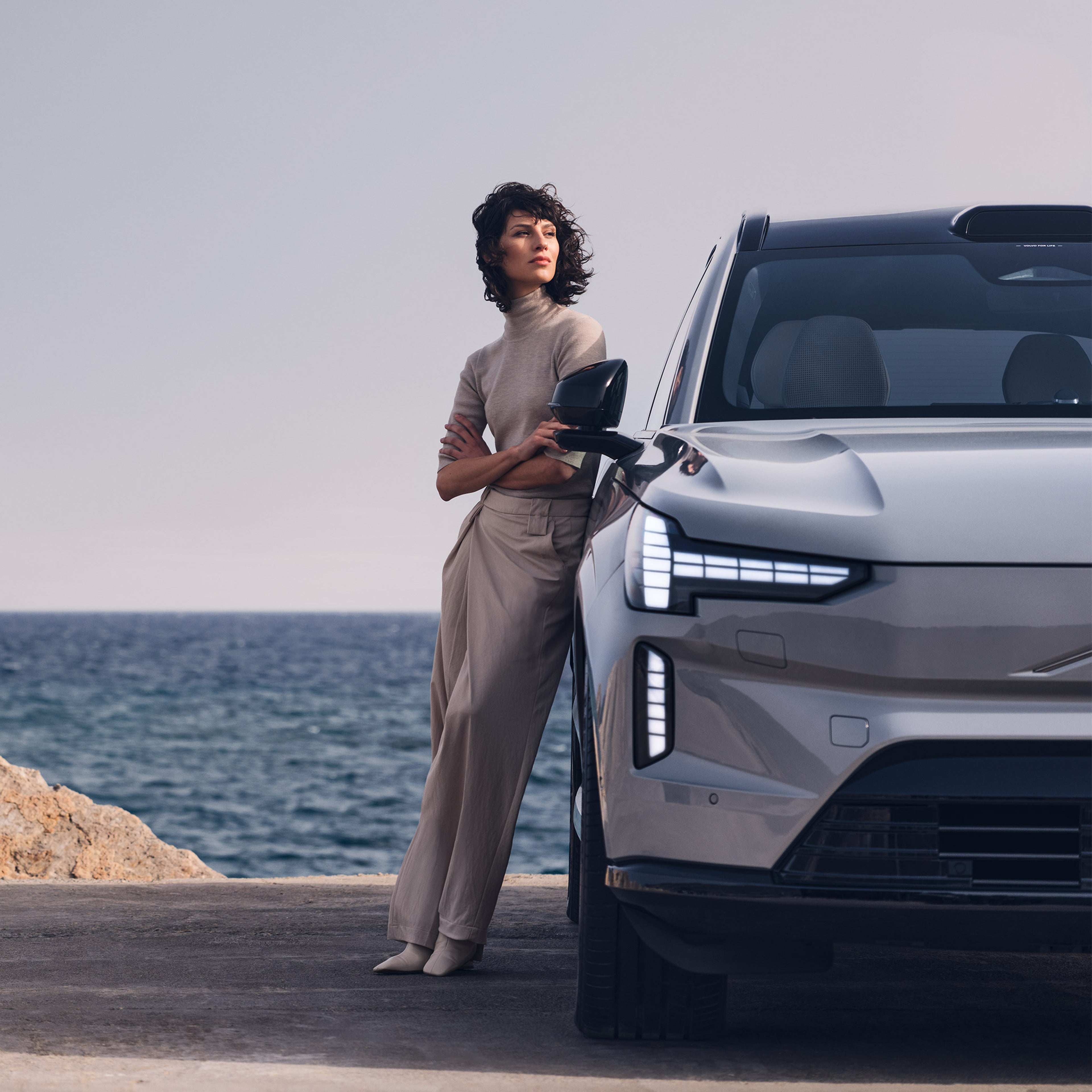 Nainen nojaa meren rantaan parkkeerattuun Vapour Grey -väriseen Volvo EX90 täyssähköautoon.