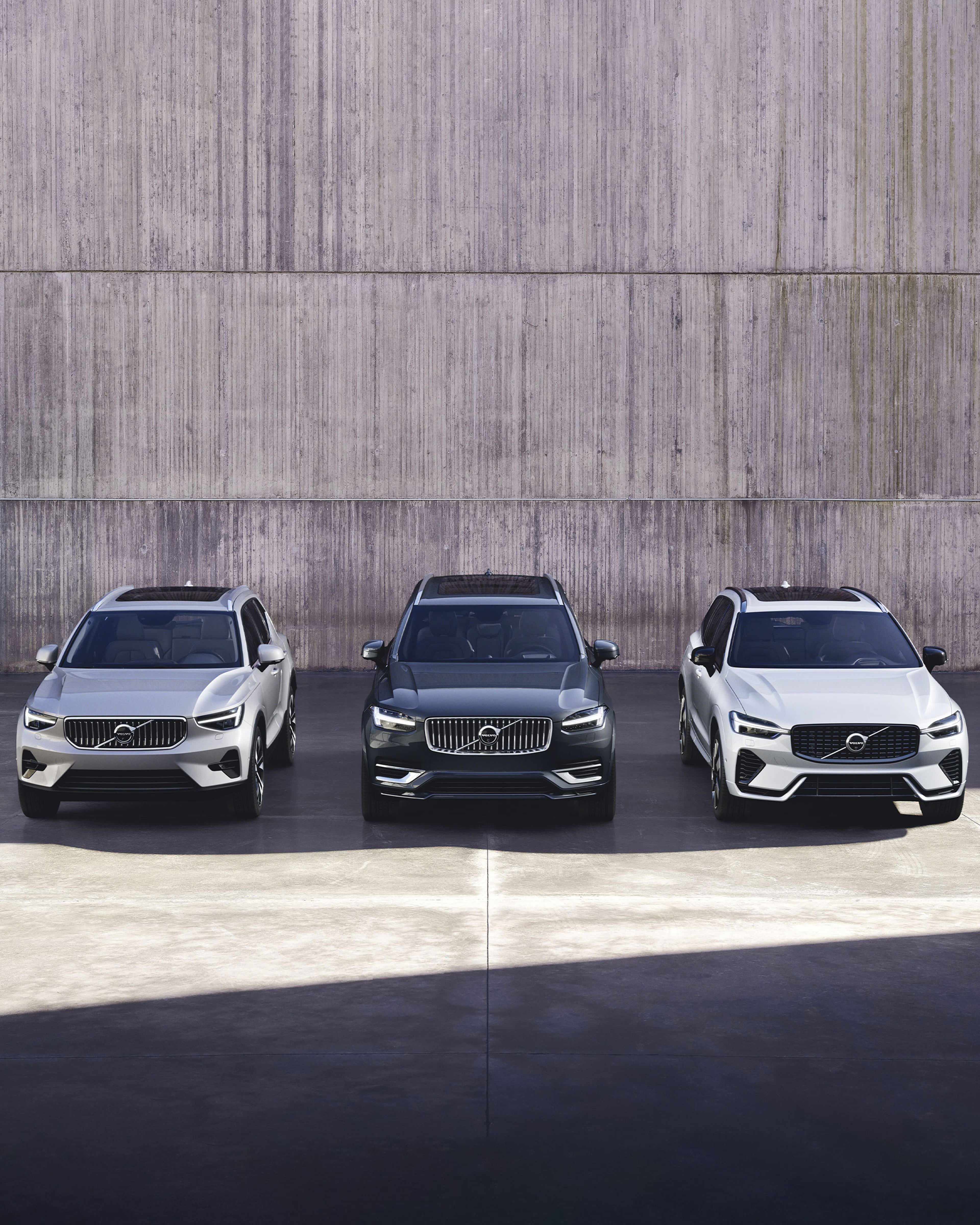 Drei Volvo Fahrzeuge in Silver Dawn (silber), Onyx Black (schwarz) und Crystal White (Weiß) stehen vor einer hellen Steinwand.