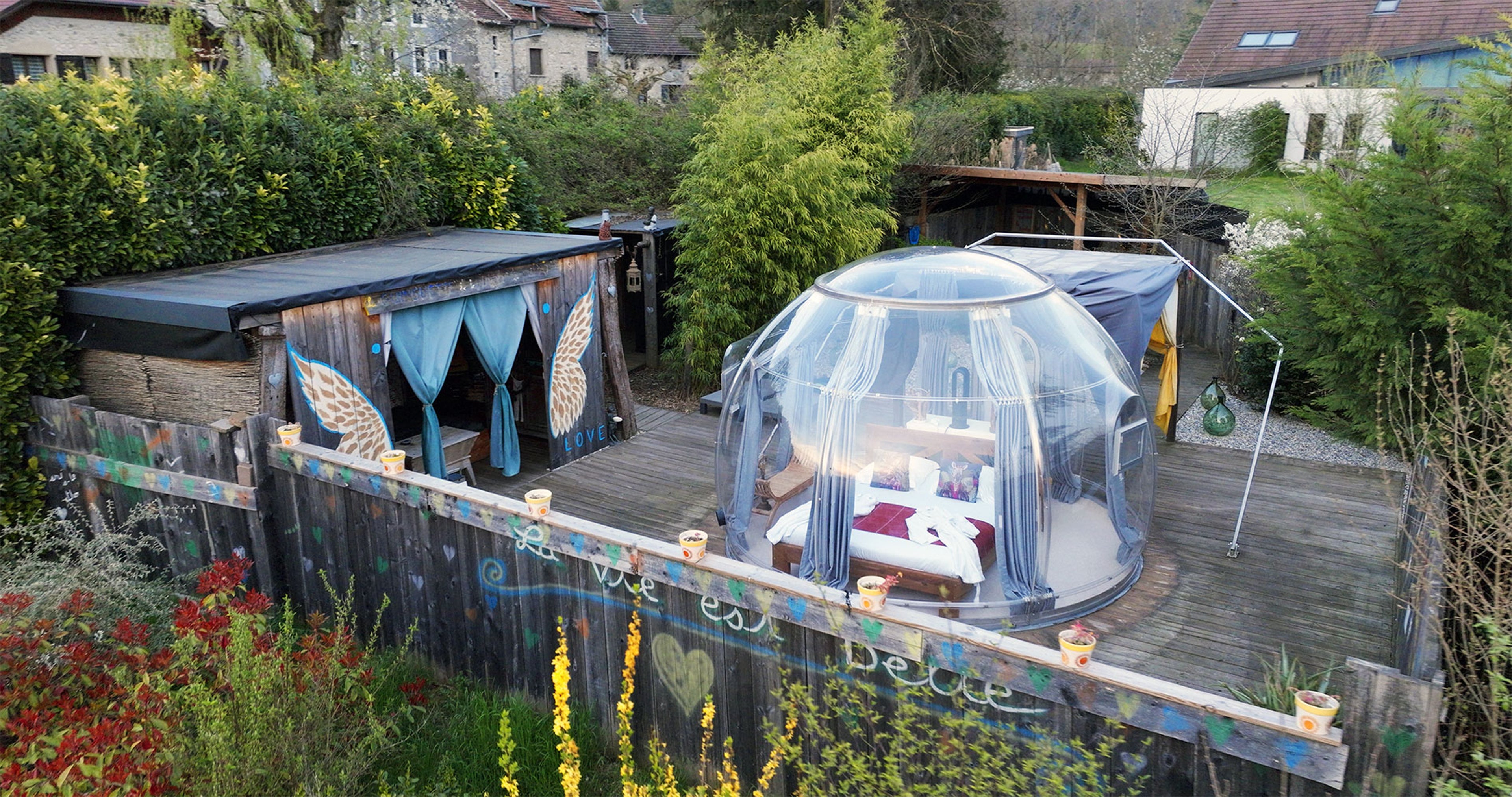 Dôme transparent avec lit sur terrasse en bois, entouré de verdure et décorations colorées.