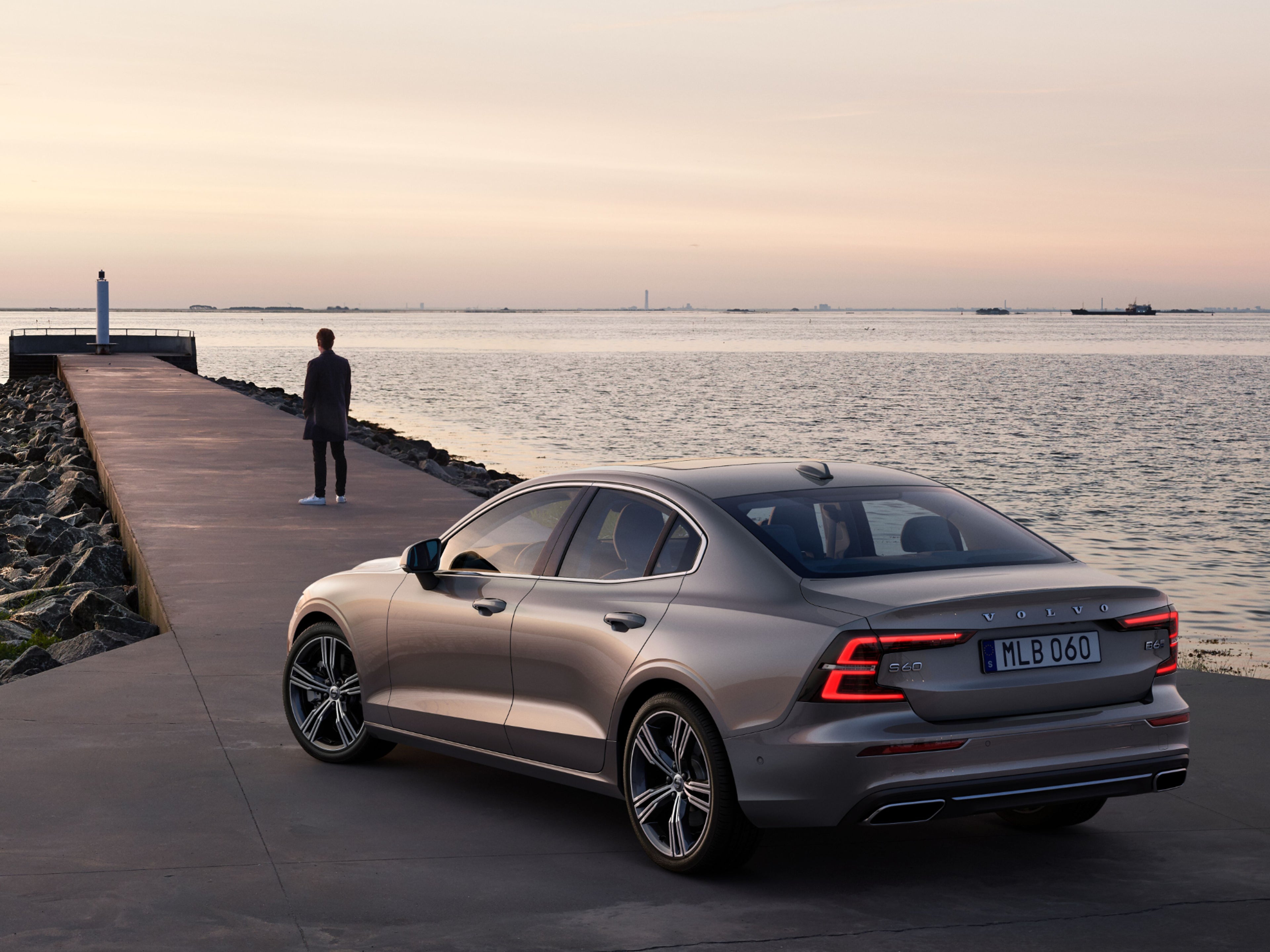 Ein Mann steht vor einer silbernen Volvo S60 Limousine auf einem Steg am Meer.