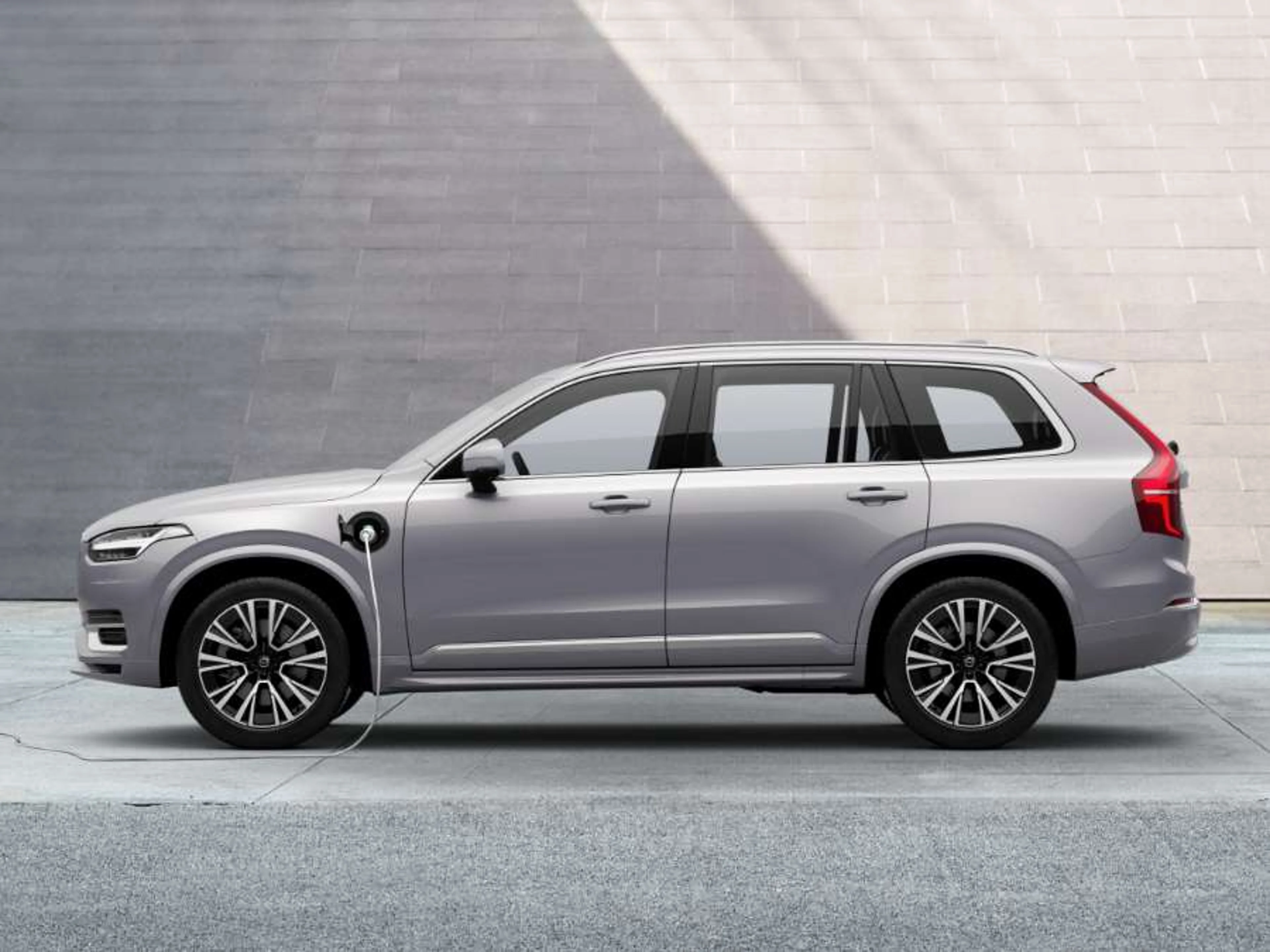 Svetlo siv Volvo XC90 plug-in hybrid v profilu, parkiran pred sodobno sivo steno med polnjenjem.