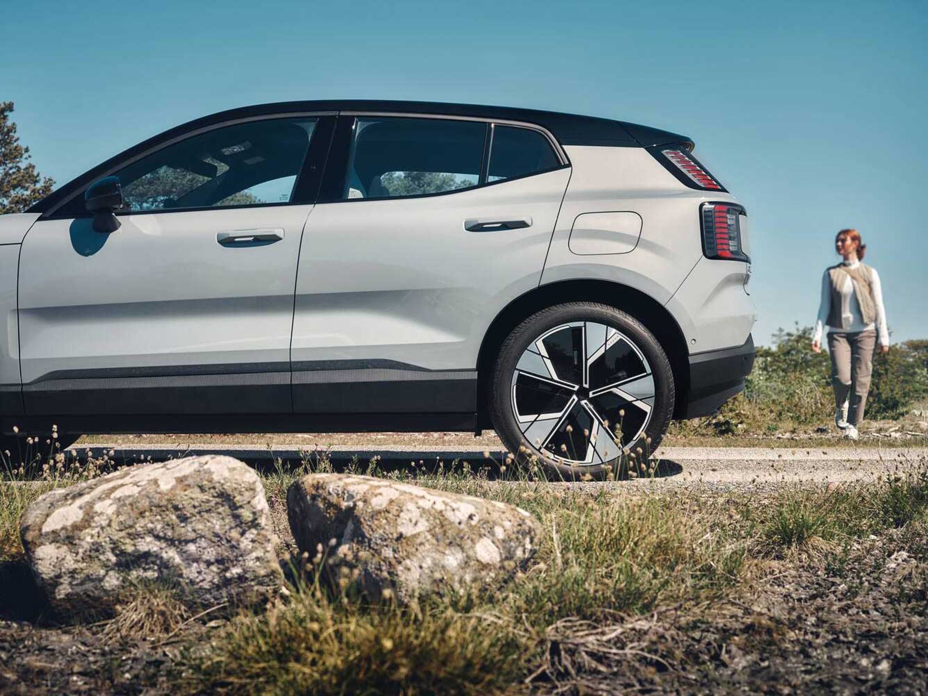 Sideview af en moderne Volvo SUV parkeret på en solrig dag. En person går på en grussti ved siden af. Nærliggende sten og græs antyder en landlig omgivelser.