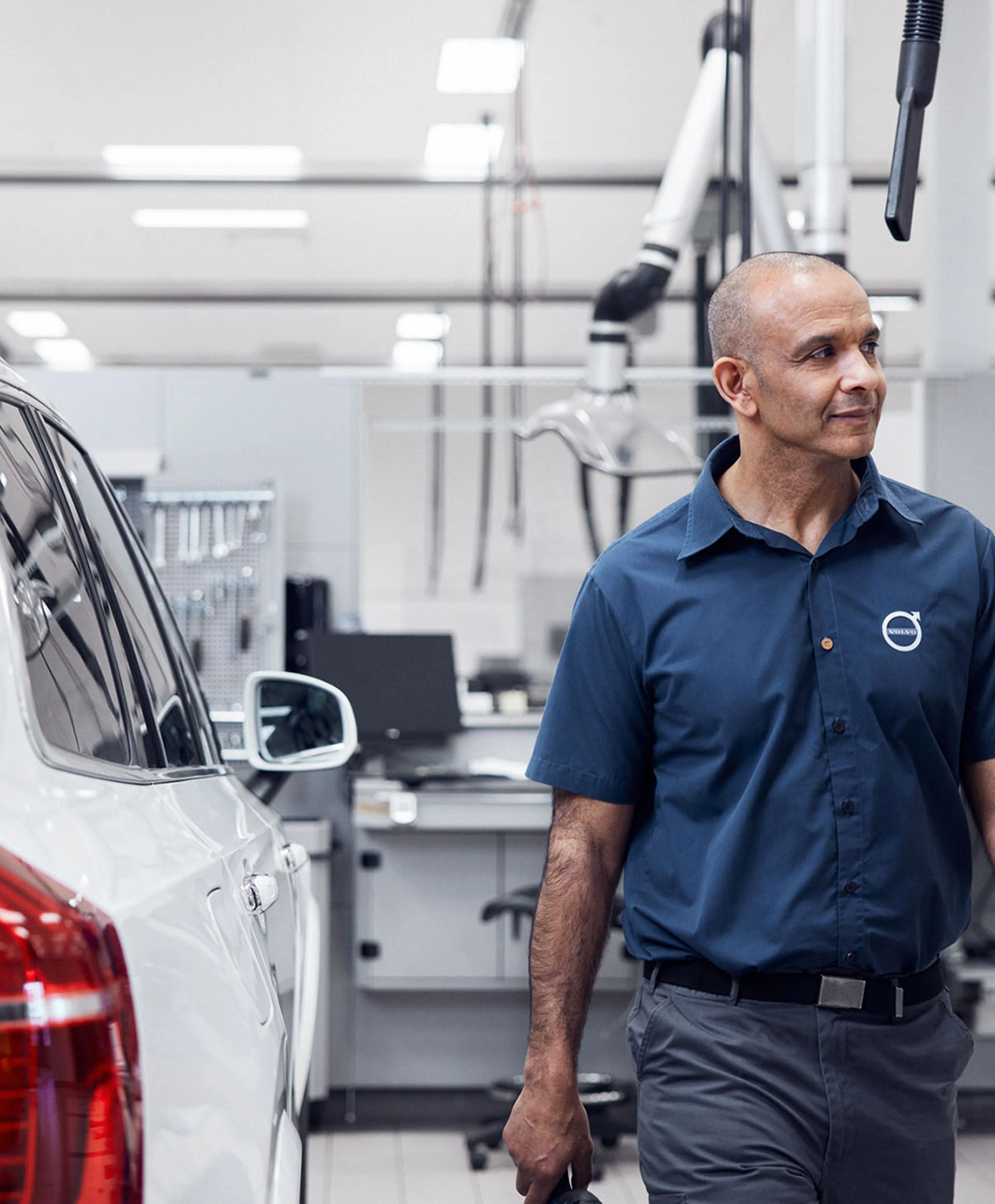 En mand i en blå arbejdstshirt står ved siden af en hvid bil i et lyst, Volvo autoværksted. Han ser tilfreds og fokuseret ud og holder et værktøj i hånden.