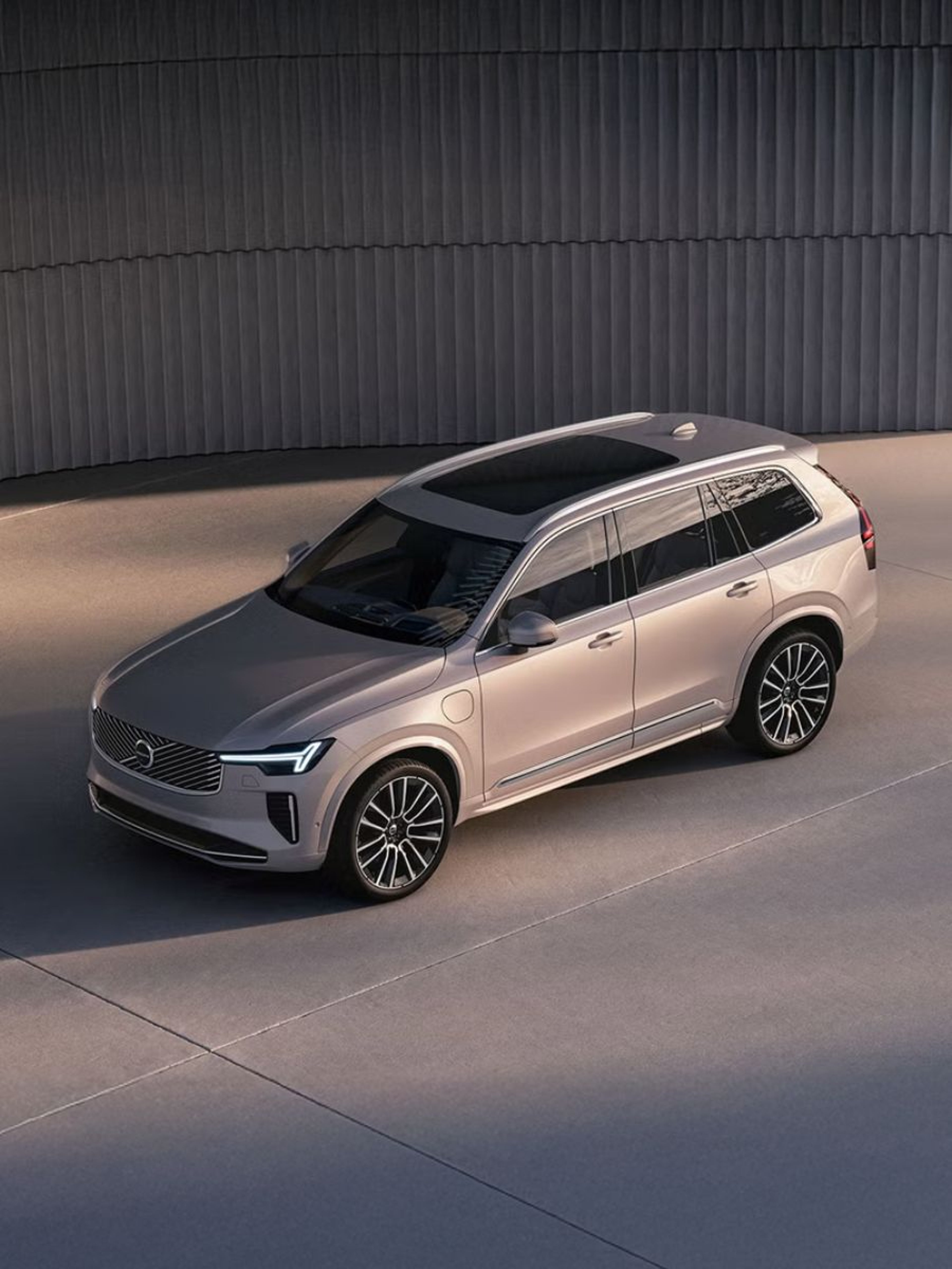 Srebrni Volvo XC90 prikazan iz zgornje perspektive, parkiran pred sodobno kovinsko fasado v studijskem okolju.