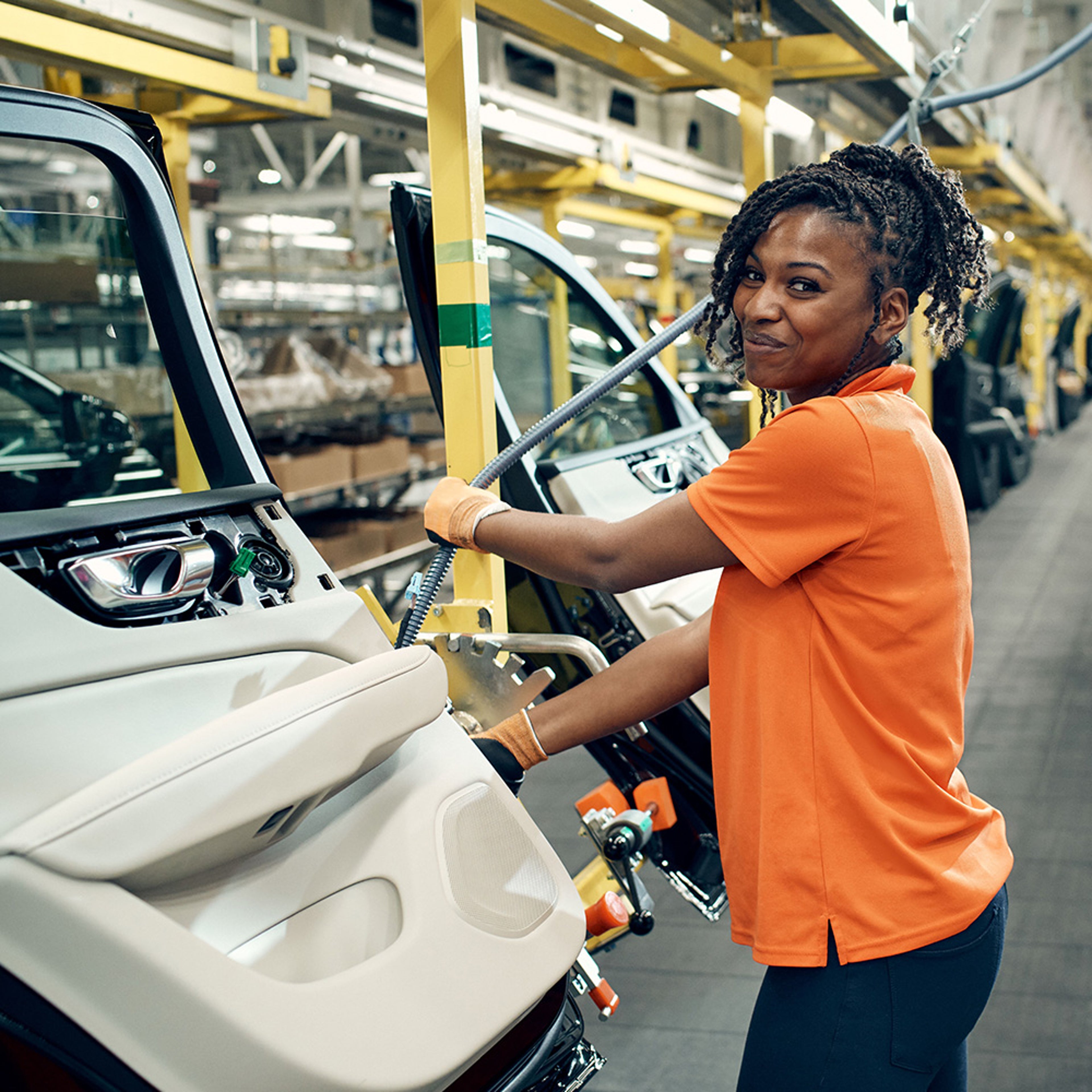 Een vrouw met een oranje shirt en werkhandschoenen werkt aan een autodeur in een Volvo fabriek.