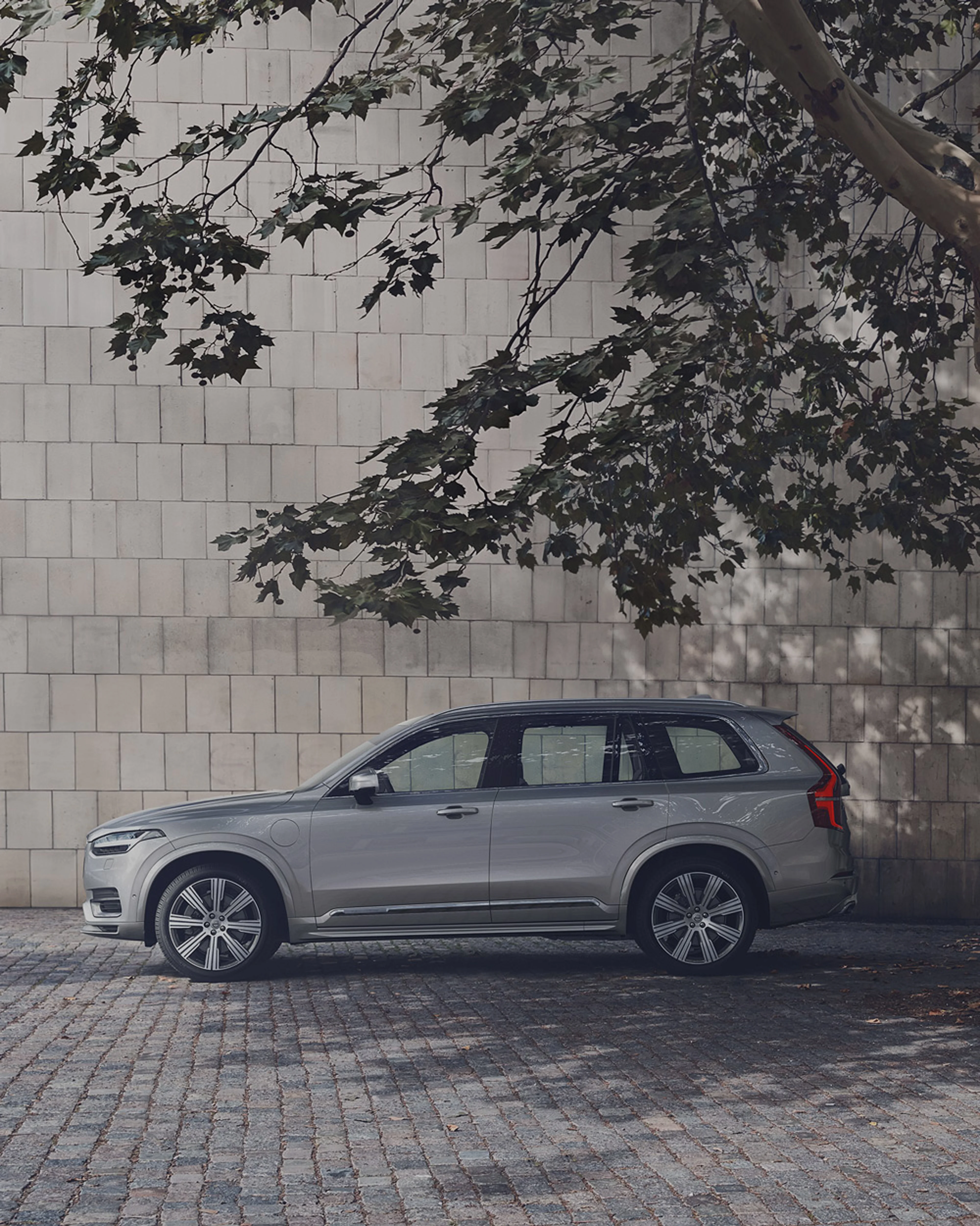 Une Volvo XC90 Recharge grise est garée à l'ombre sous un arbre dans une cour.