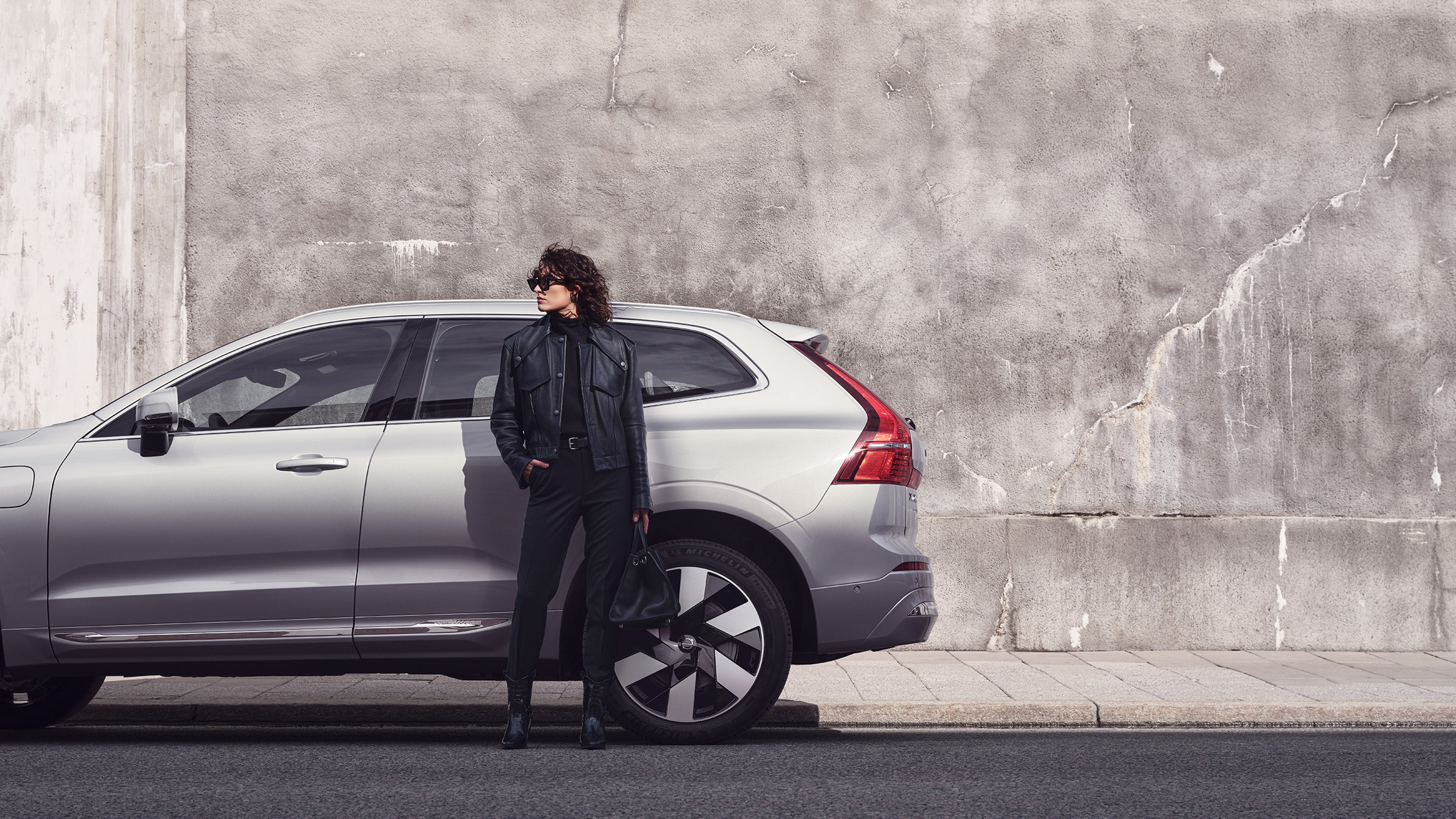 Eine schwarz gekleidete Frau steht mit der Hand in der Tasche vor einem grauen Volvo XC60 und schaut zur Seite.