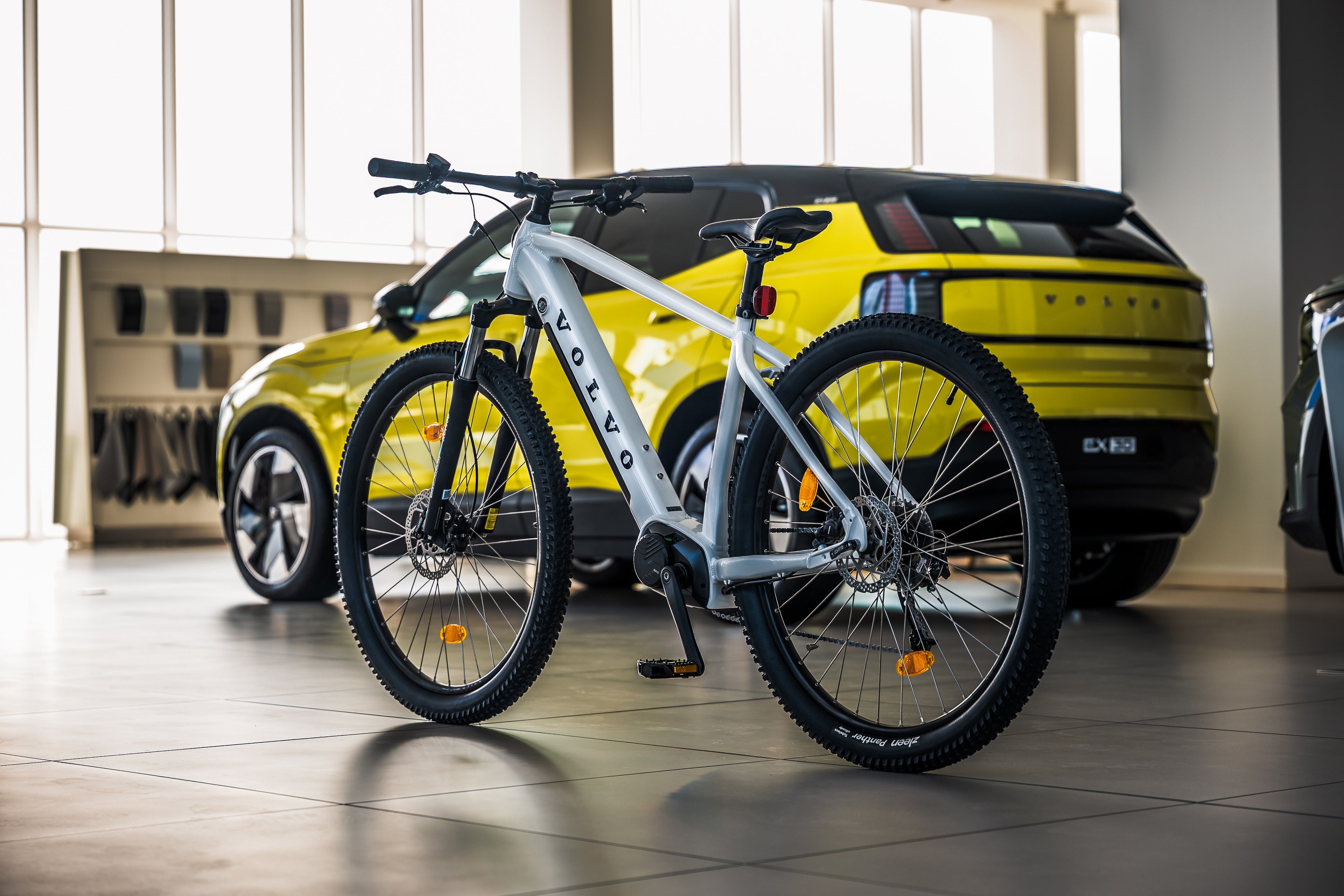 Eлектрически велосипед в бял цвят стои изправен на фона на Volvo EX30 в цвят Moss Yellow