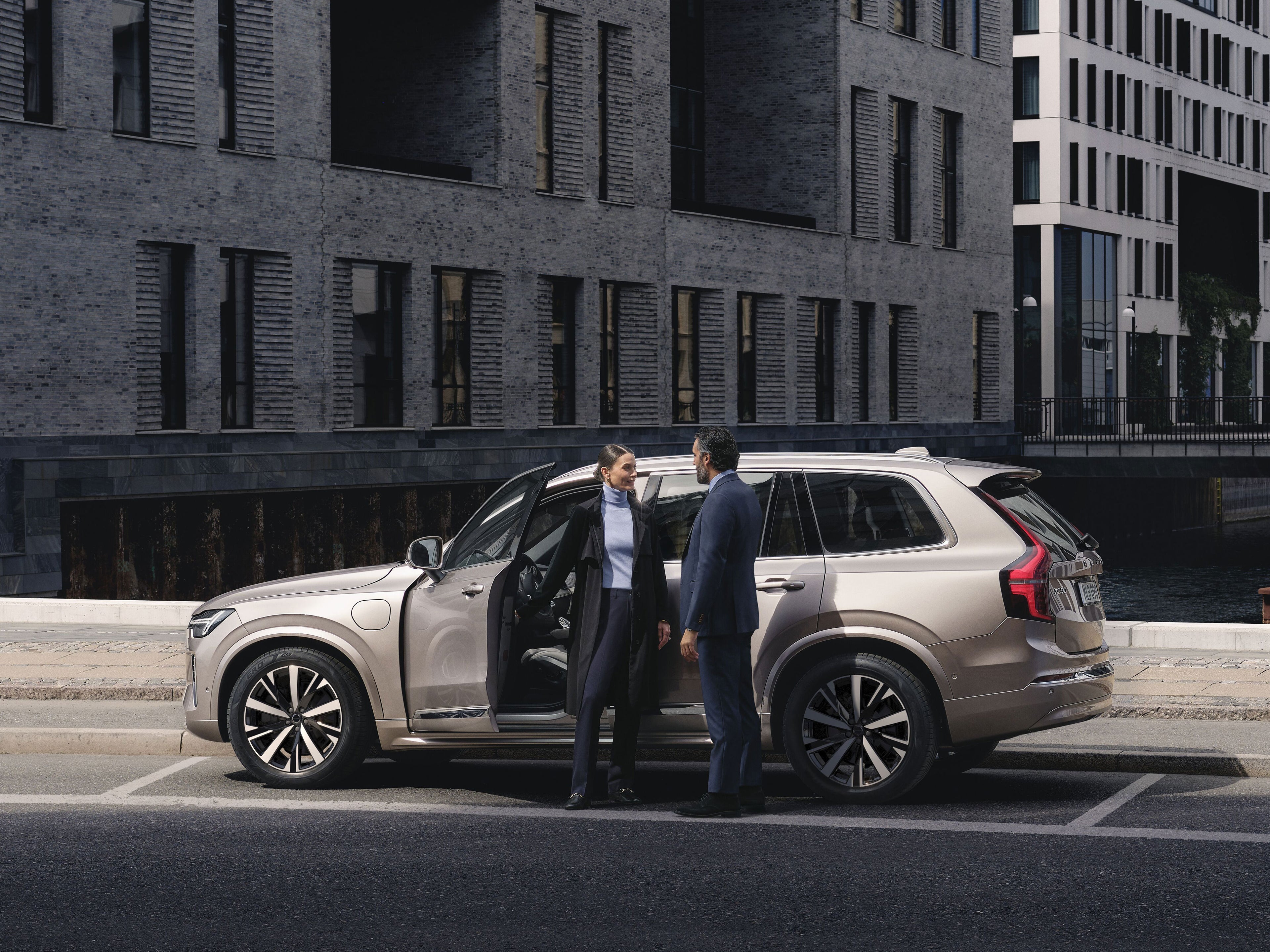 Eine Frau und ein Mann stehen an einem beig-silbernen Volvo XC90 SUV und unterhalten sich.