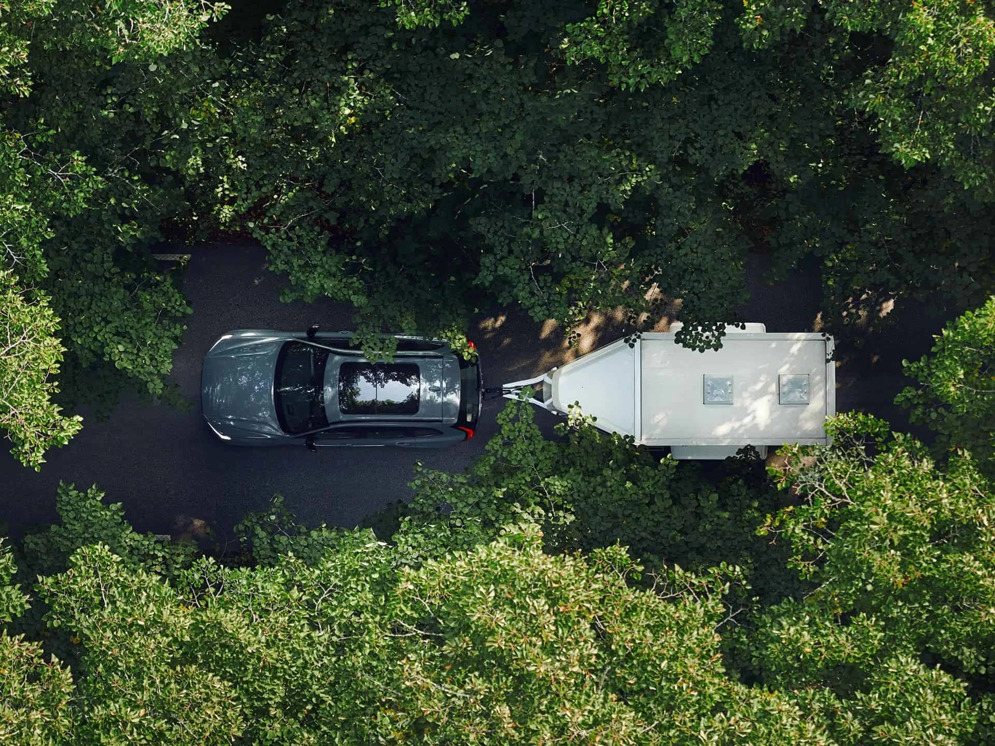 Bovenaanzicht van een grijze auto met een witte caravan, rijdend op een weg door het bos