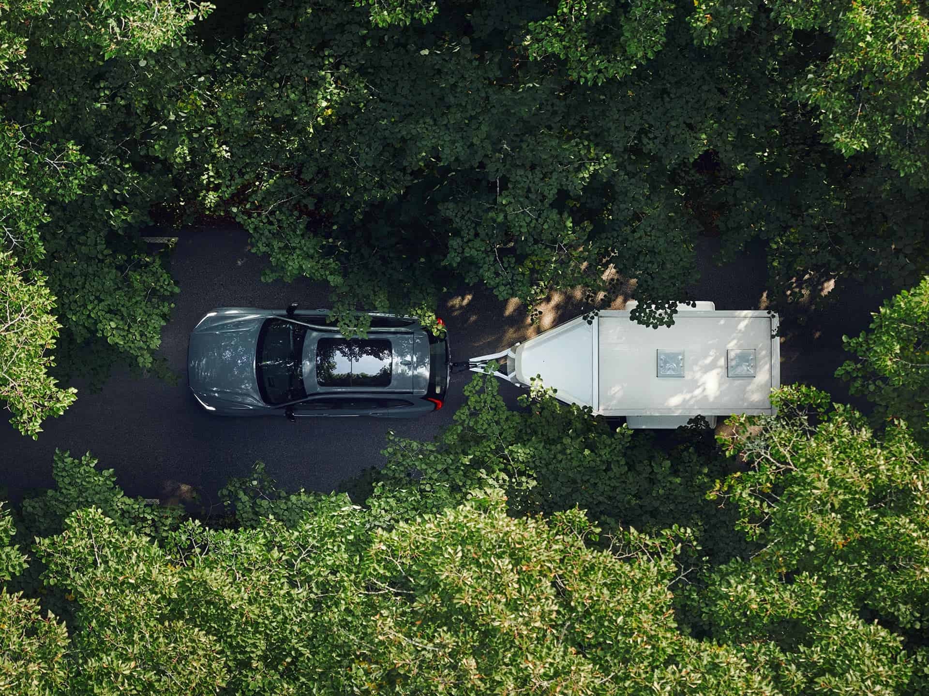 Une Volvo grise tracte une caravane blanche sur une route à travers une forêt, vue de dessus.