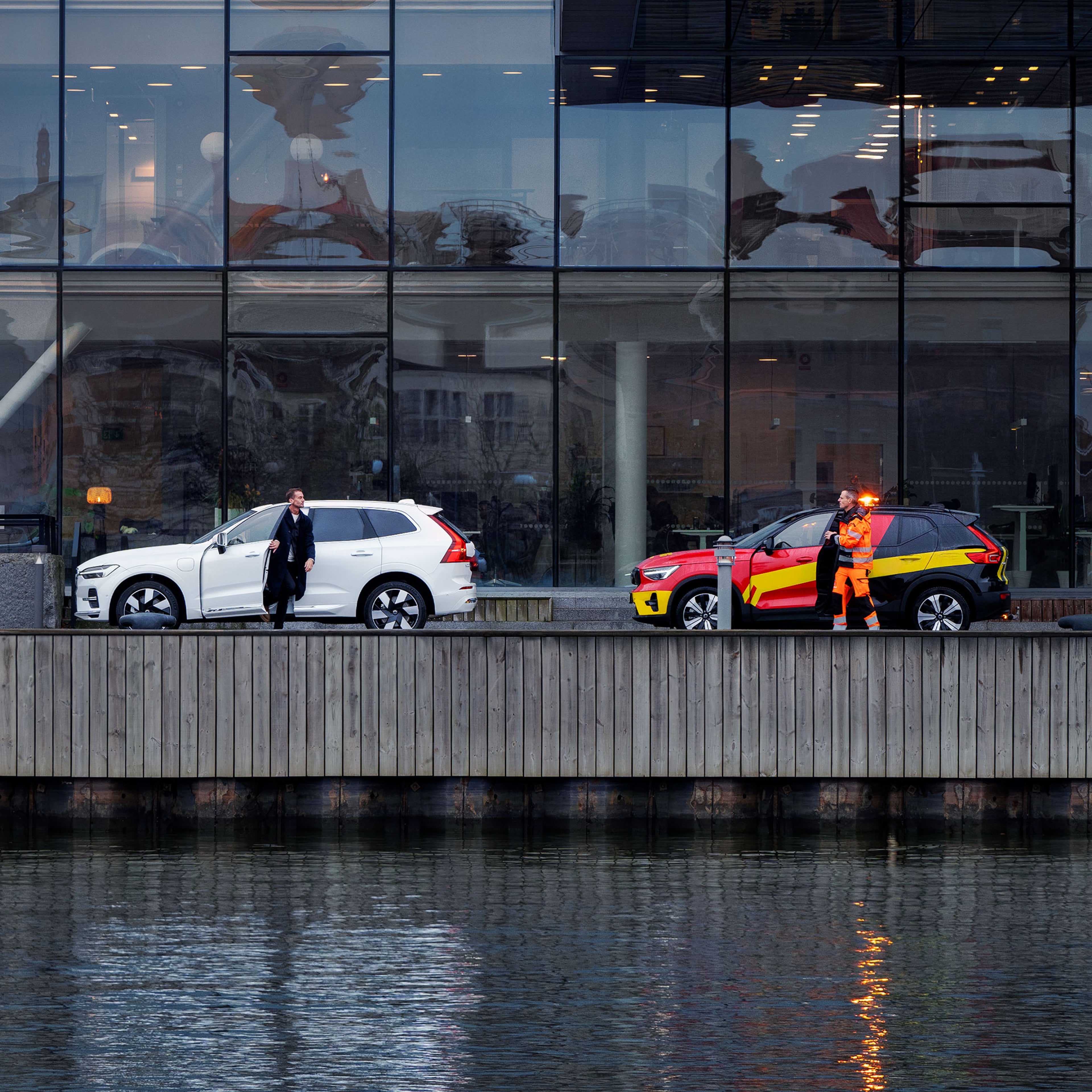 Mies tulee ulos rantakadulle pysäköidystä valkoisesta Volvo -autosta. Perään on pysäköity Volvo Tiepalvelun auto, jonka kuski on noussut autosta. Taustalla on toimistorakennus.