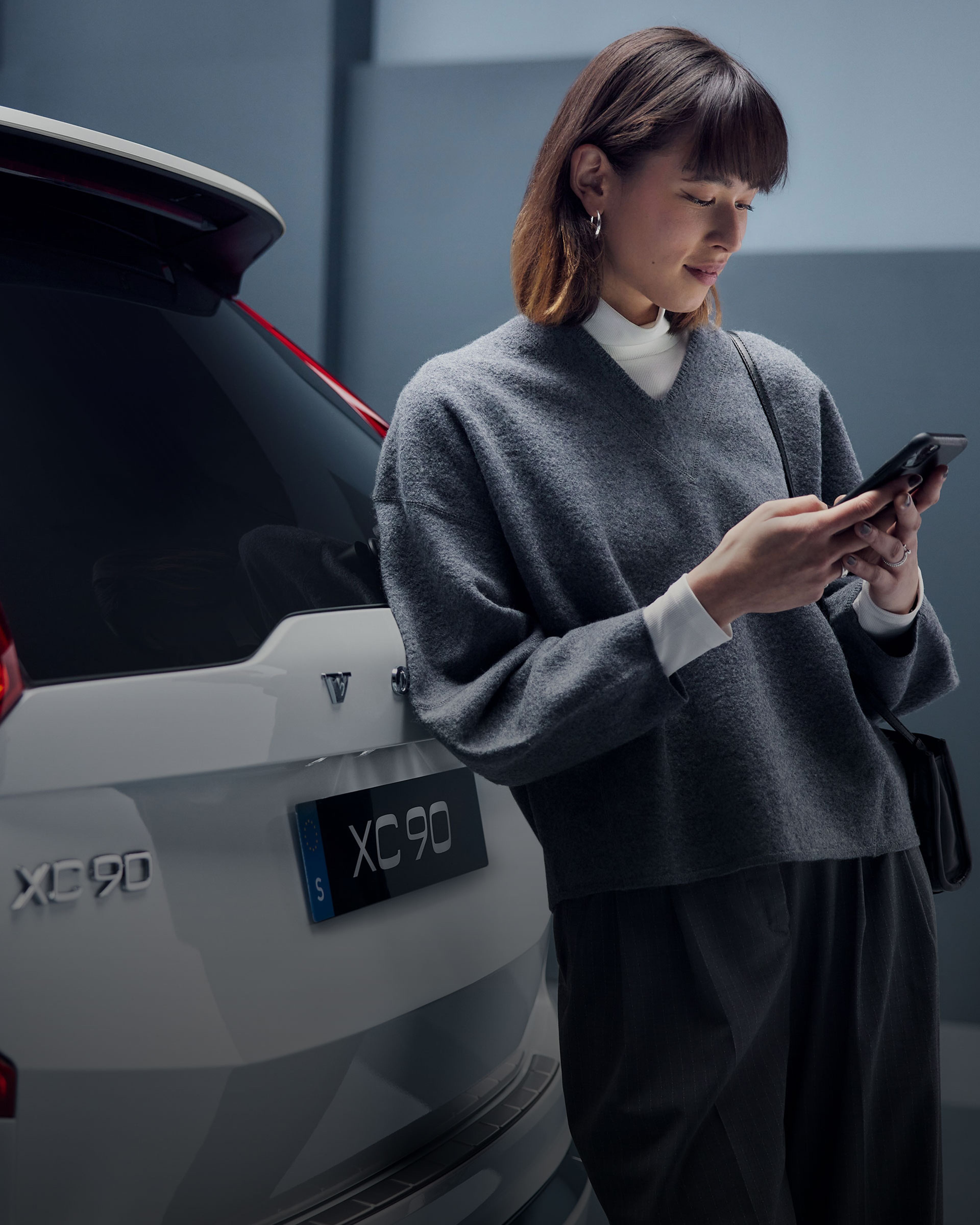 Una mujer recargada sobre un Volvo XC90 con su teléfono en la mano.
