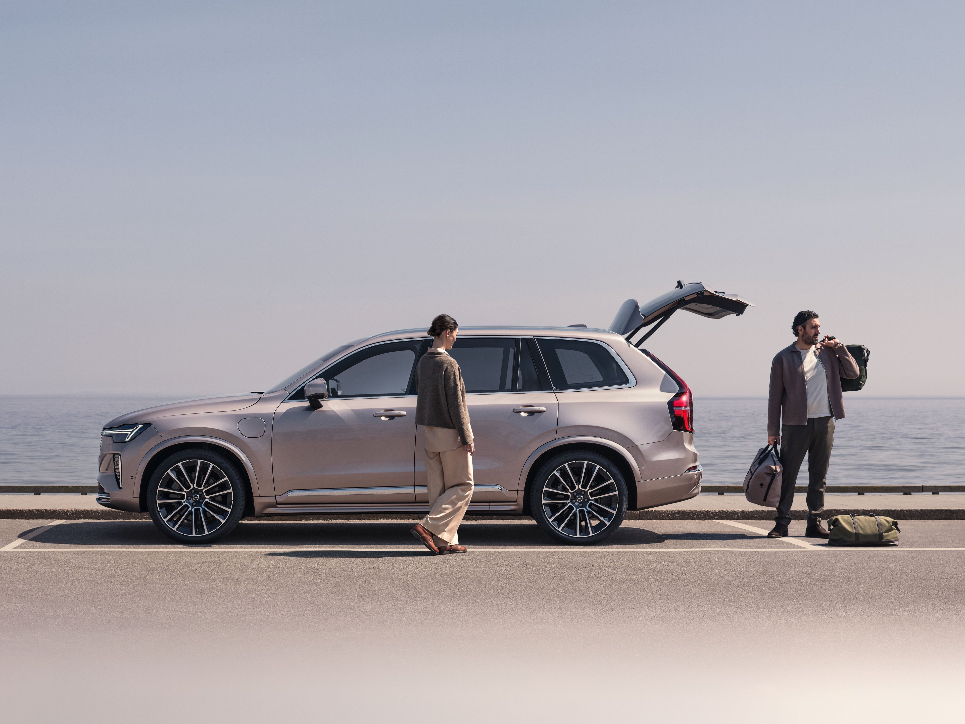 Zwei Personen stehen an einem silbernen Volvo XC90 SUV, der in der Nähe eines Sees steht.