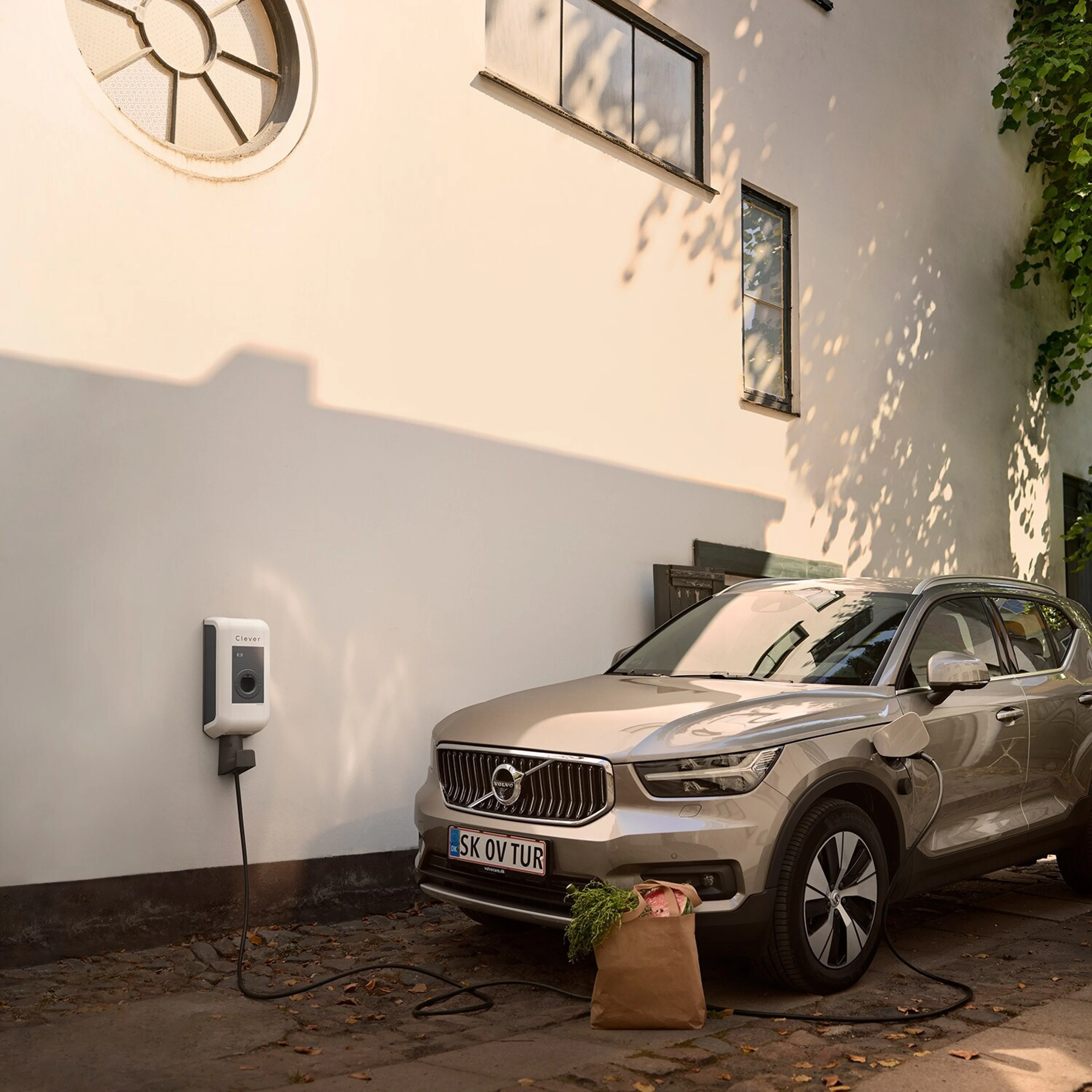 Sølvfarvet elektrisk Volvo SUV lader op ved en vægmonteret ladestation udenfor en hvid bygning. Solbeskinnet scene med skygger og en papirspose med dagligvarer i nærheden.