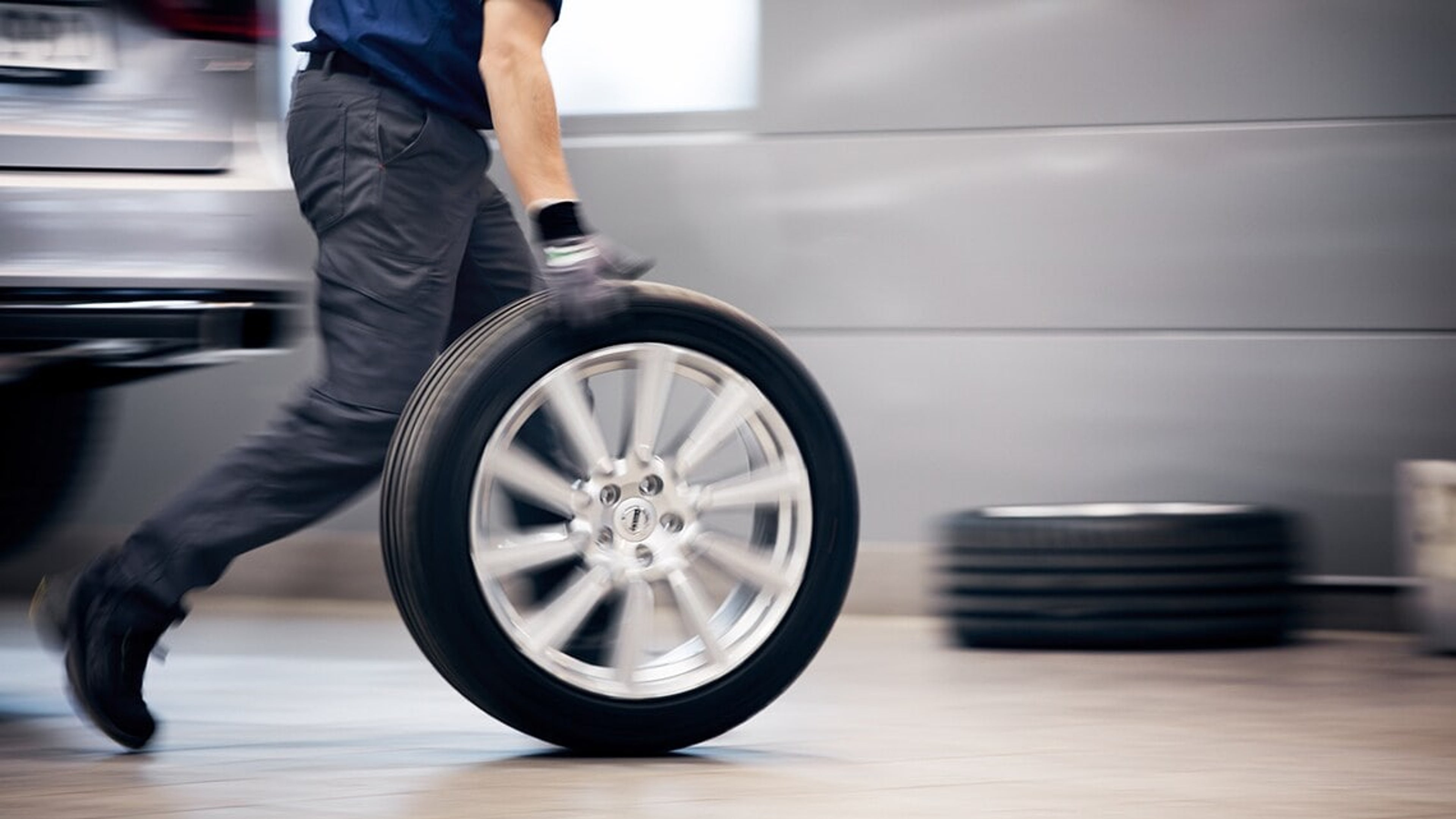 En Volvo mekaniker i bevægelse iført blå skjorte og handsker ruller et bildæk i et værksted, hvilket udstråler handling og effektivitet. En bil og en stak dæk ses sløret i baggrunden.
