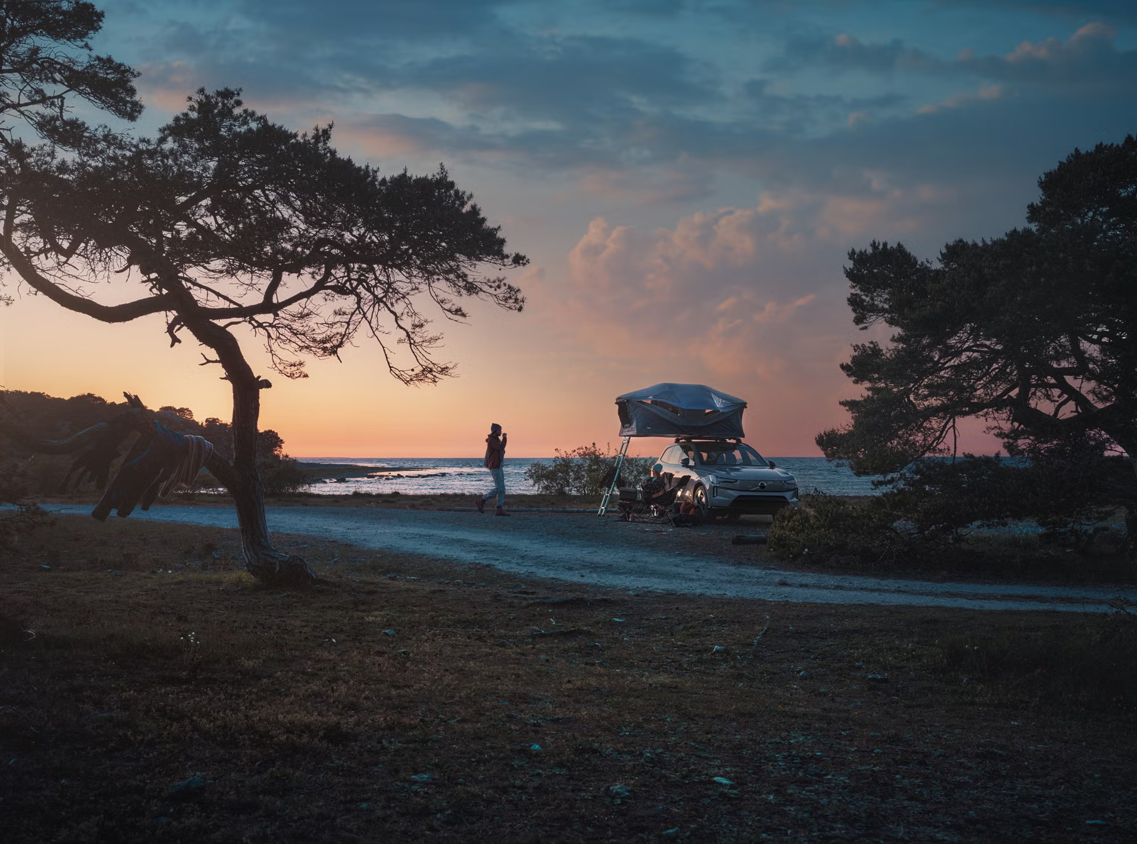 Ein grauer Volvo steht mit einem Volvo Dachzelt auf dem Dach vor einem Meer. Im Hintergrund geht rot orange die Sonne unter.