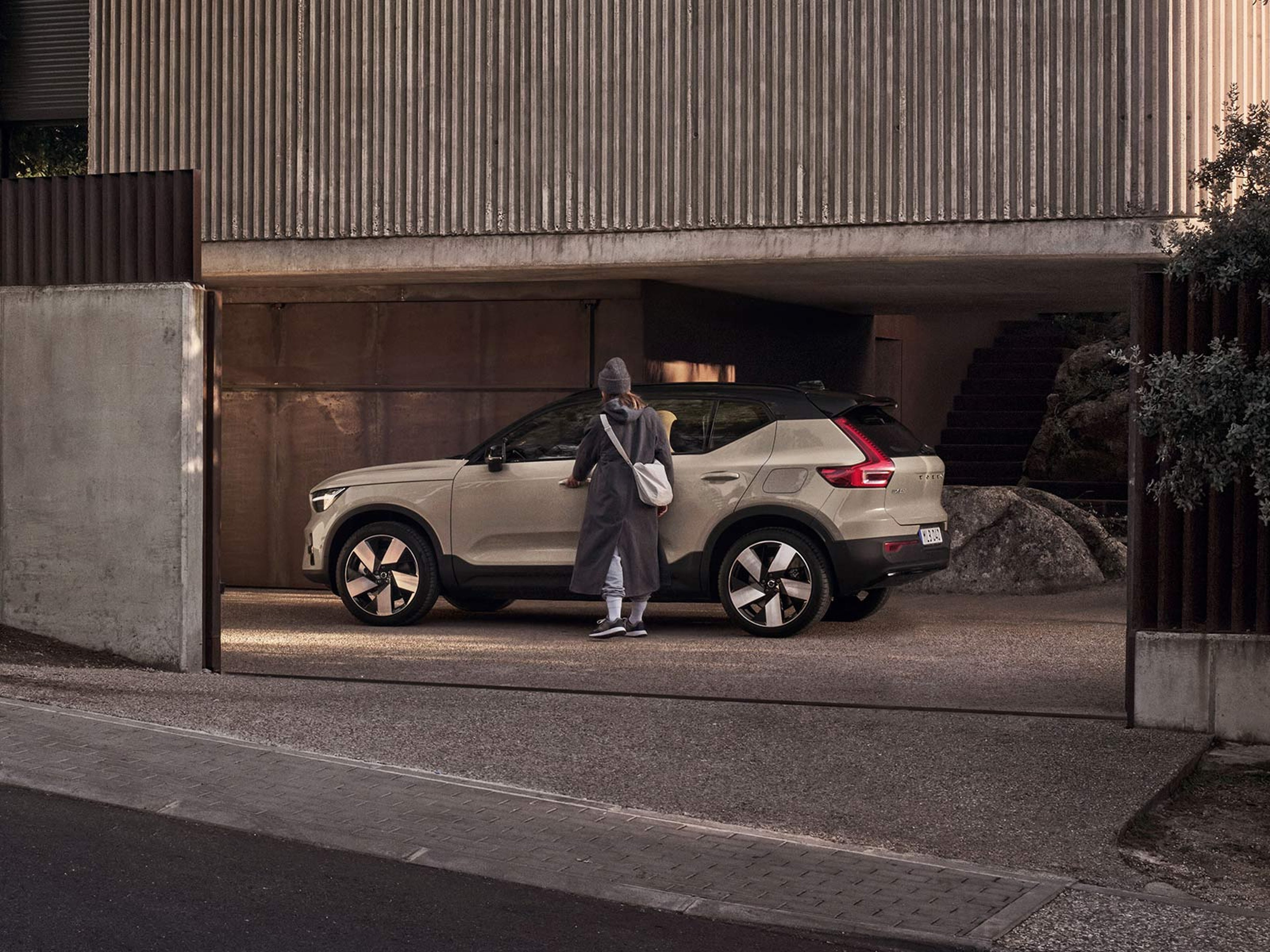 Beige EX40 parkerad framför ett hus i sten, en kvinna i grå kappa ska gå in i bilen.