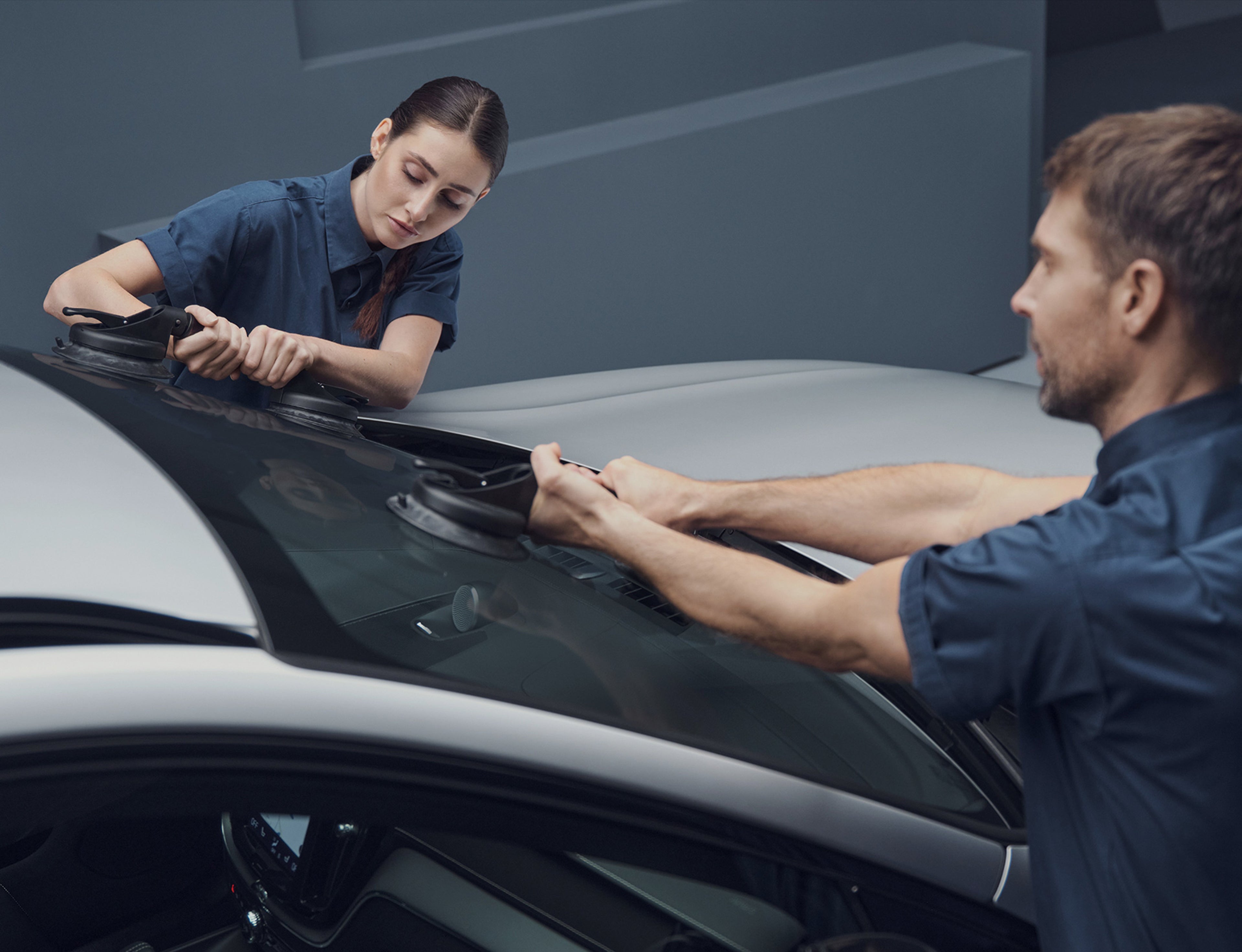 Deux techniciens Volvo remplacent le pare-brise d'une Volvo argentée dans un atelier Volvo à l'aide de ventouses.