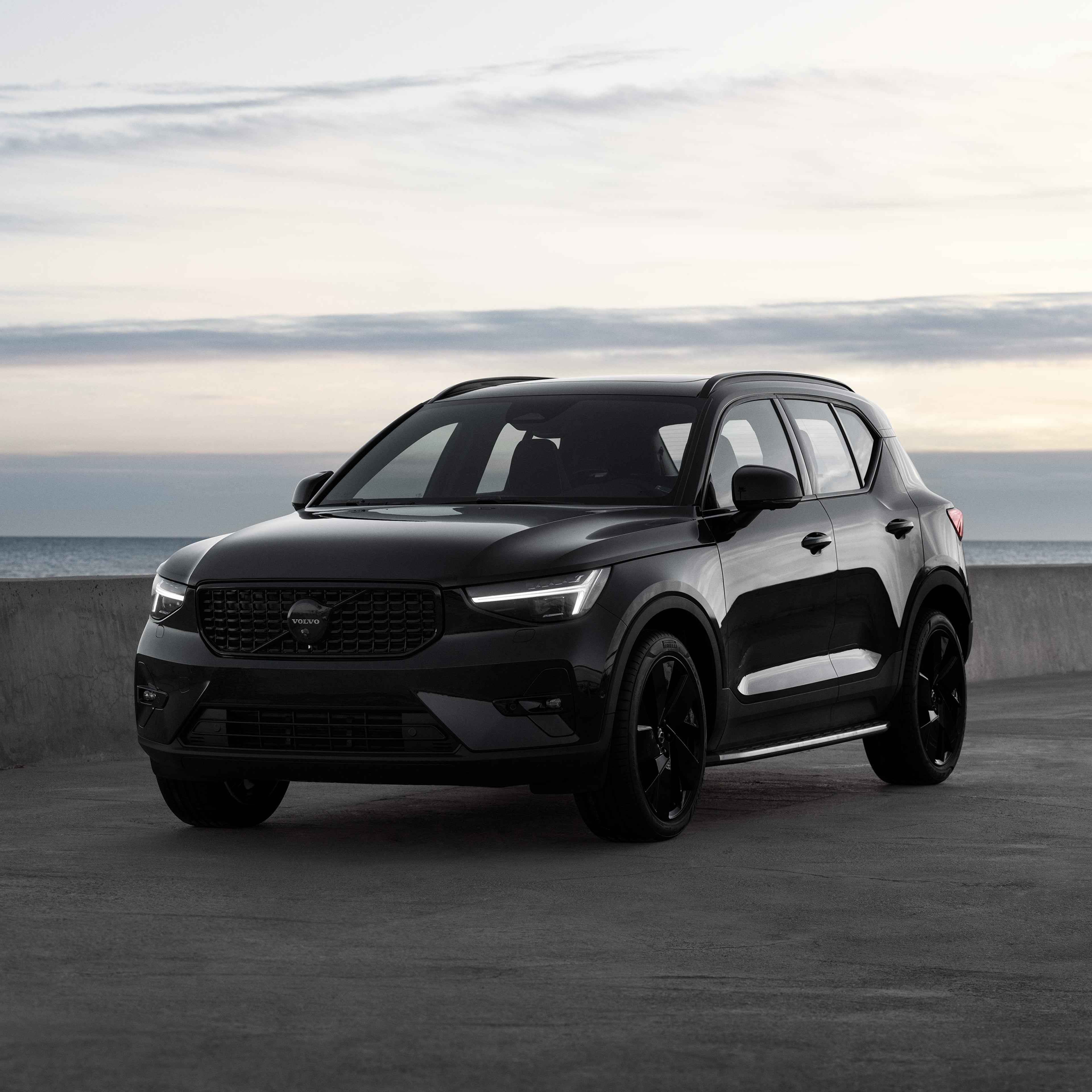 Volvo XC40 Black Edition kevythybridiauto pysäköitynä betonitasolle. Taustalla meri ja auringon valaisema taivas, jossa on hentoja valkoisia pilviä.