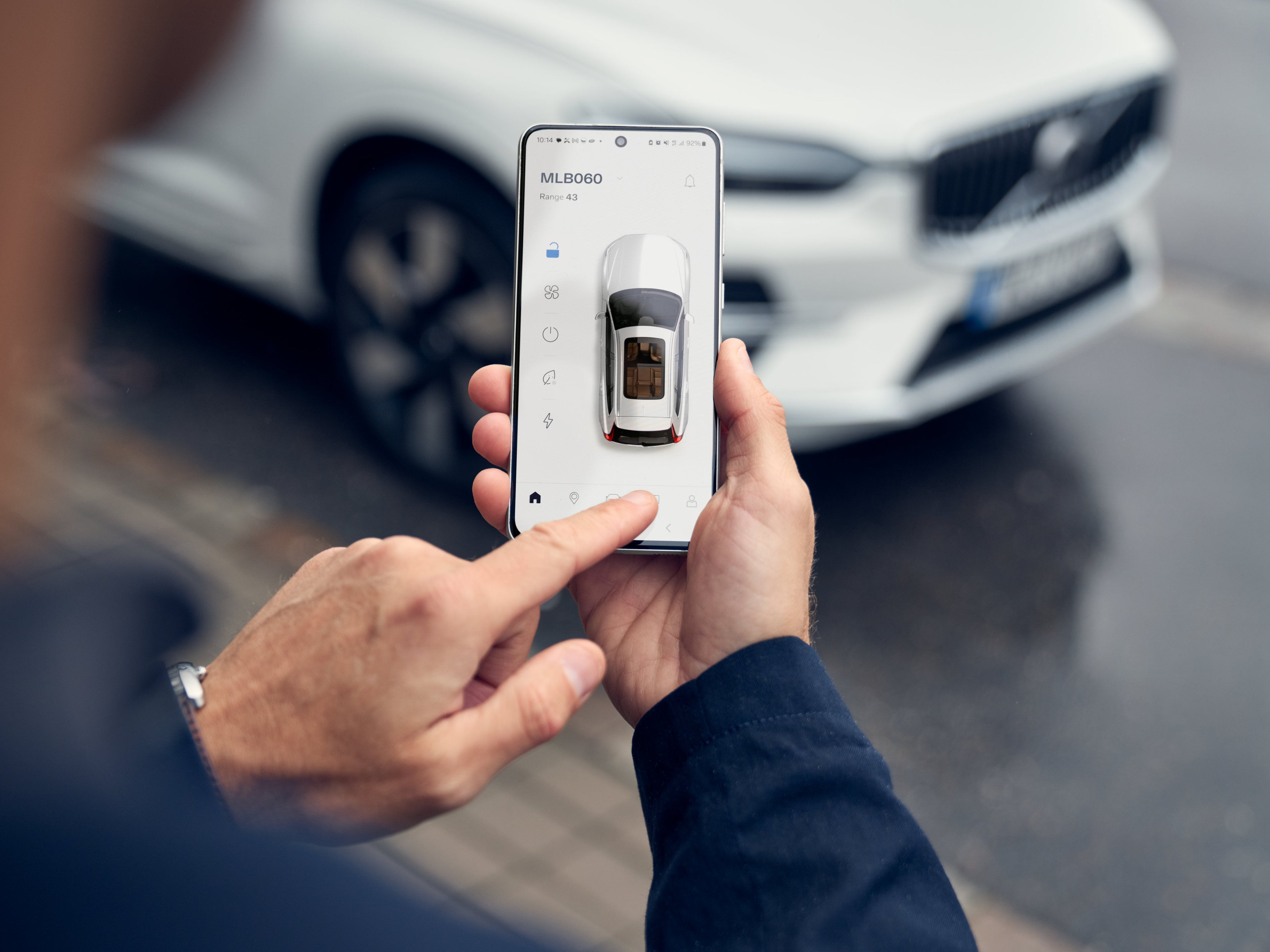 Eine rechts Hand hält ein Handy in der Hand und die linke Hand zeigt mit dem Zeigefinger auf das Display des Handys. Auf dem Bildschirm ist ein Fahrzeug in der Volvo Cars App abgebildet.