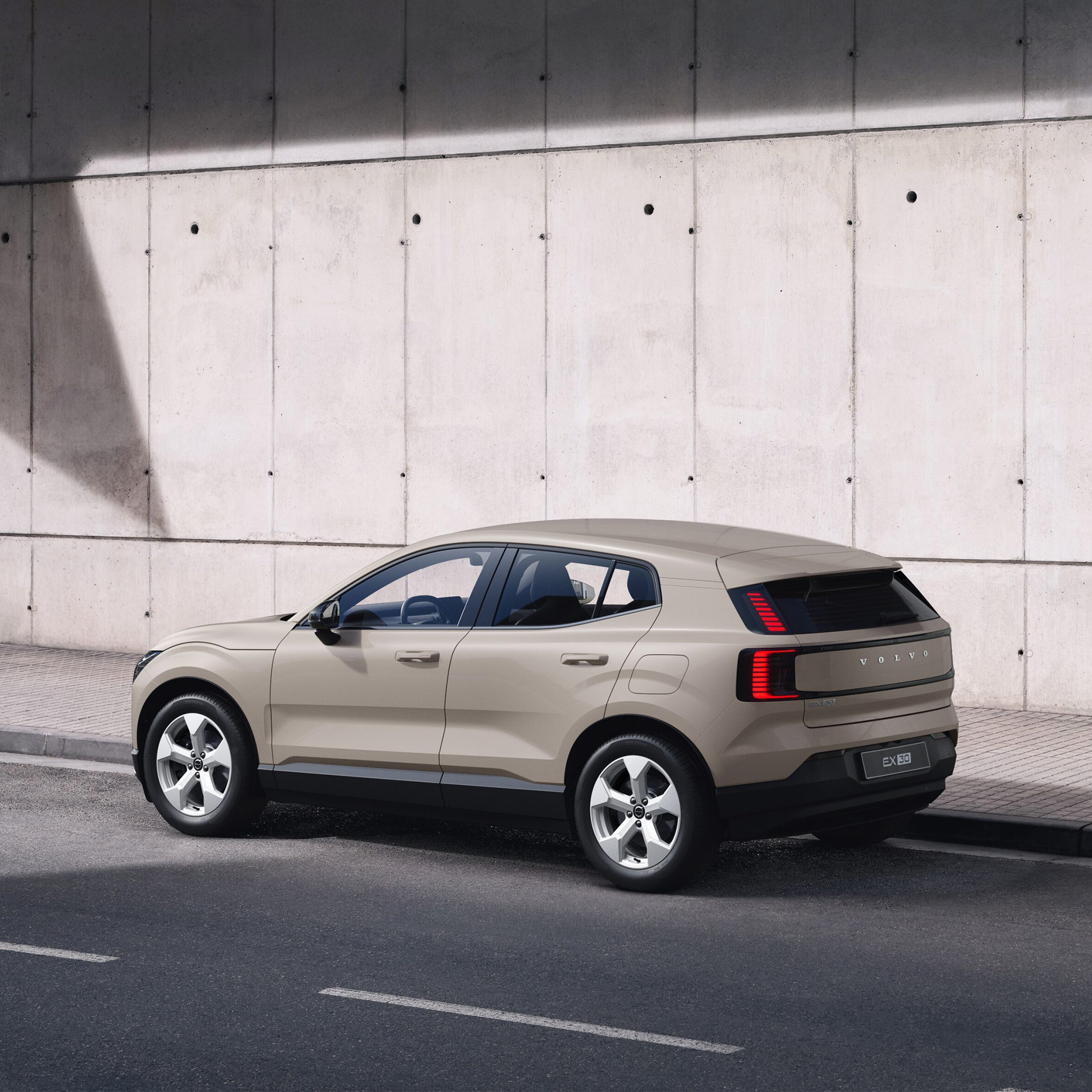 Sand dune -värinen Volvo EX30 Essential täyssähköinen katumaasturi pysäköitynä kadun varteen. Taustalla kivetty jalkakäytävä ja betoniseinä.
