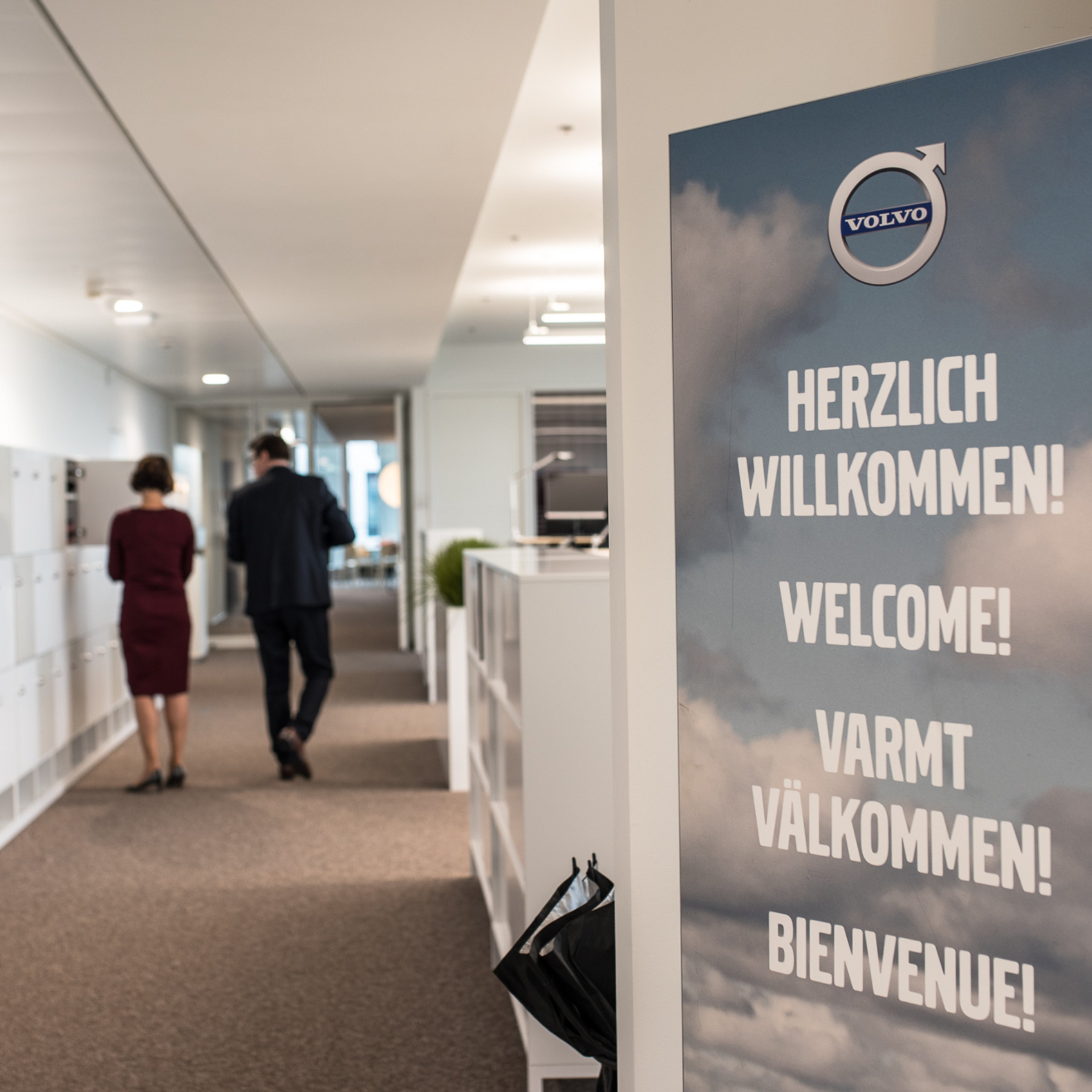 Im Fokus ist ein Willkommensschild von Volvo in der "Herzlich Willkommen" in verschiedenen Sprachen steht und im Hintergrund erkennt man einen Büro Gang mit zwei Personen die sich unterhalten.